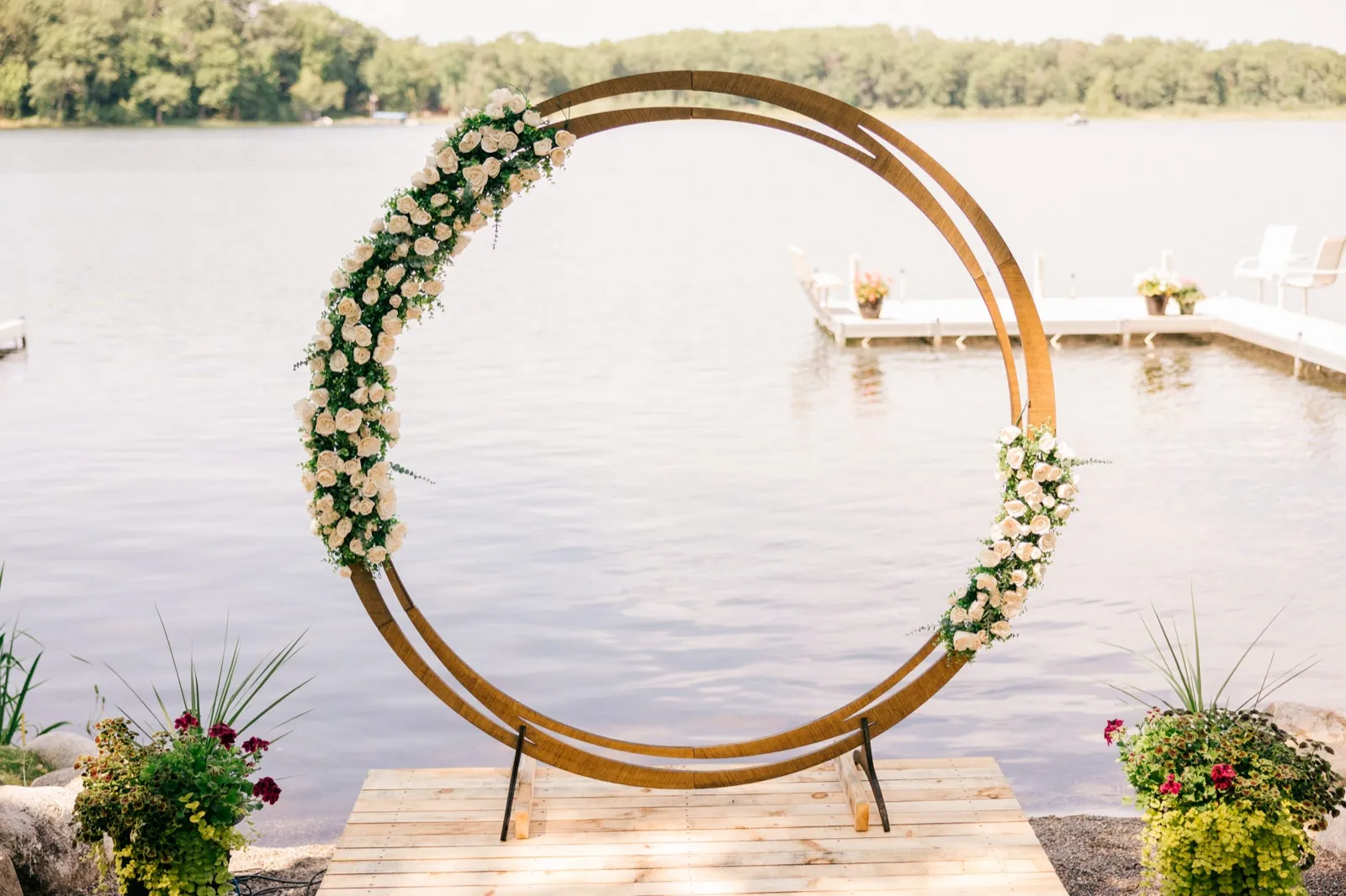 A circular wooden ceremony arch wrapped with white roses and greenery stands on a wooden platform at the edge of a lake, with a dock and trees visible across the water. — Tim Larsen Photography, Brainerd Lakes MN