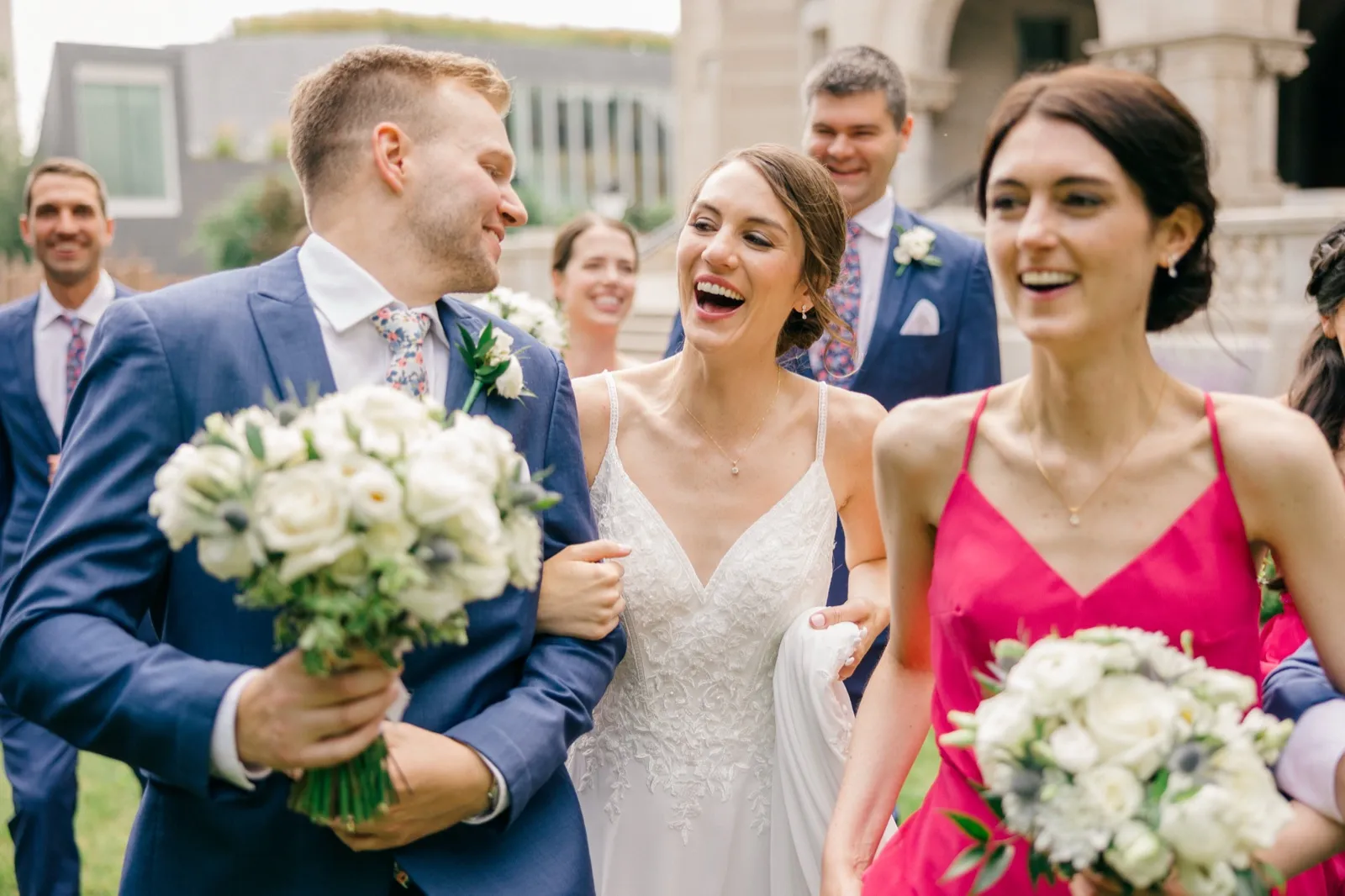 The bride laughs openly while walking arm-in-arm with the groom and bridal party across the lawn, surrounded by bouquets and smiling friends. — Tim Larsen Photography, Minneapolis MN