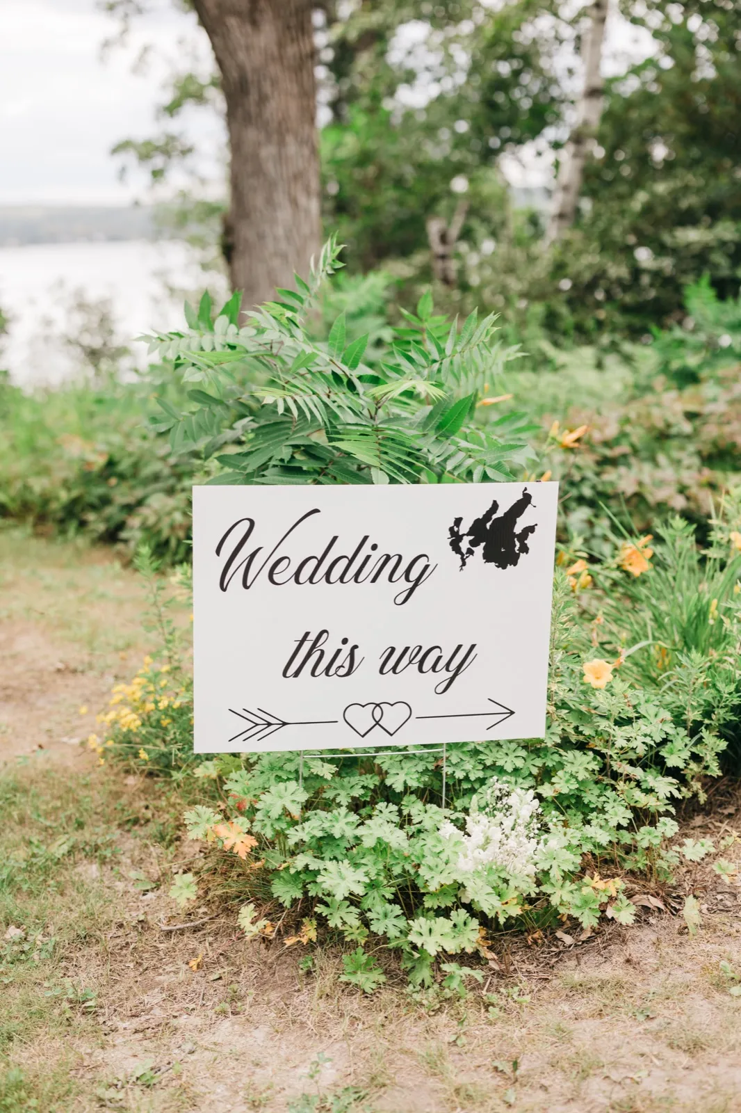 A hand-lettered Wedding This Way sign stands among ferns and greenery near the lake at Chase on the Lake. — Tim Larsen Photography, Brainerd Lakes MN
