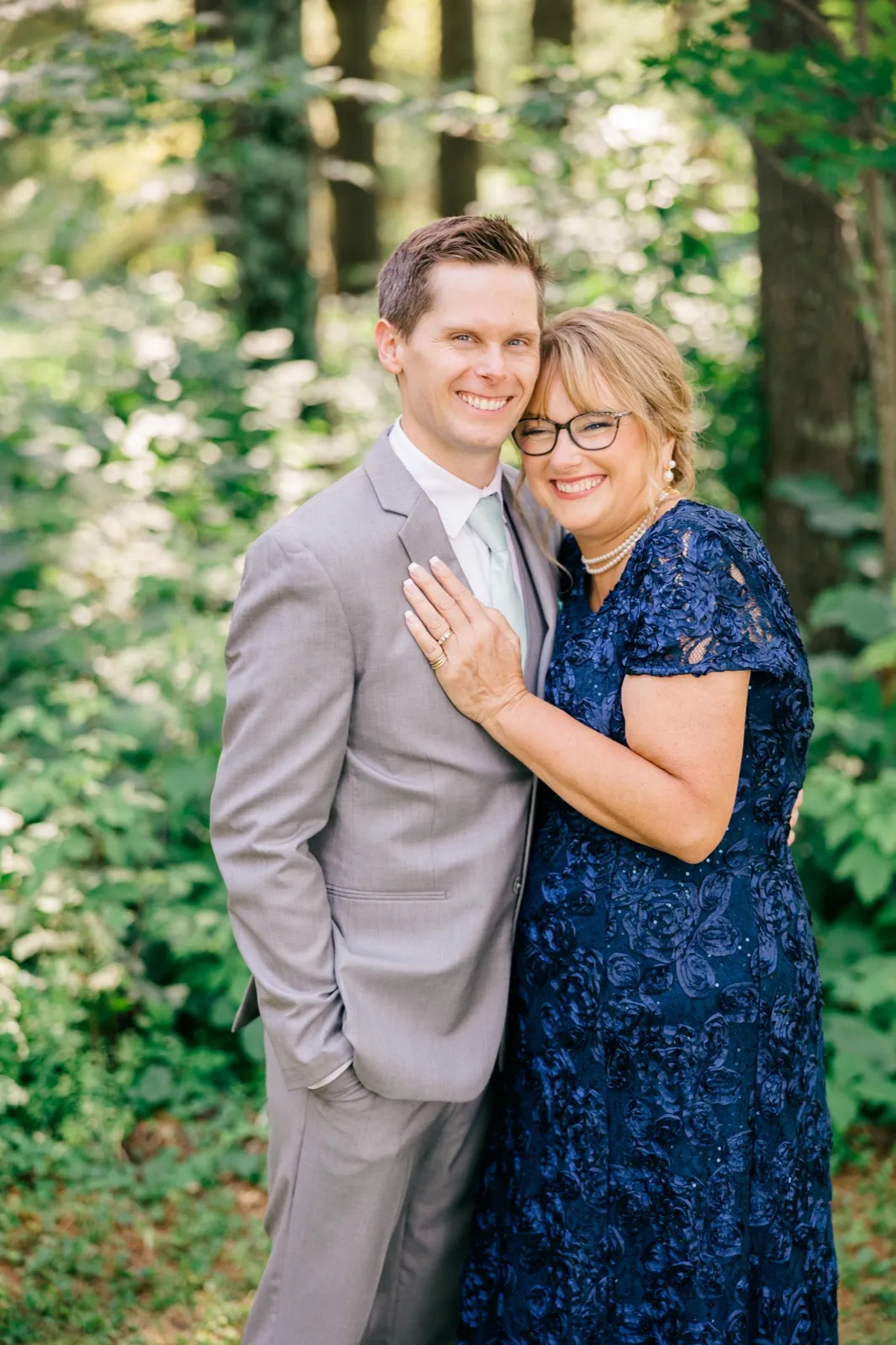 The groom in a gray suit smiles beside a woman in a navy floral dress with pearls as they embrace in a sunlit forest. — Tim Larsen Photography, Brainerd Lakes MN