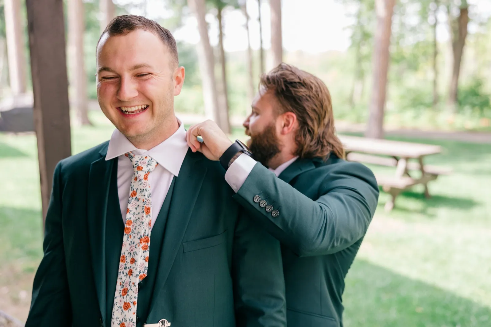 Eric laughs with his eyes closed in a green suit and floral tie while a groomsman behind him adjusts his jacket lapel — Tim Larsen Photography, Brainerd Lakes MN