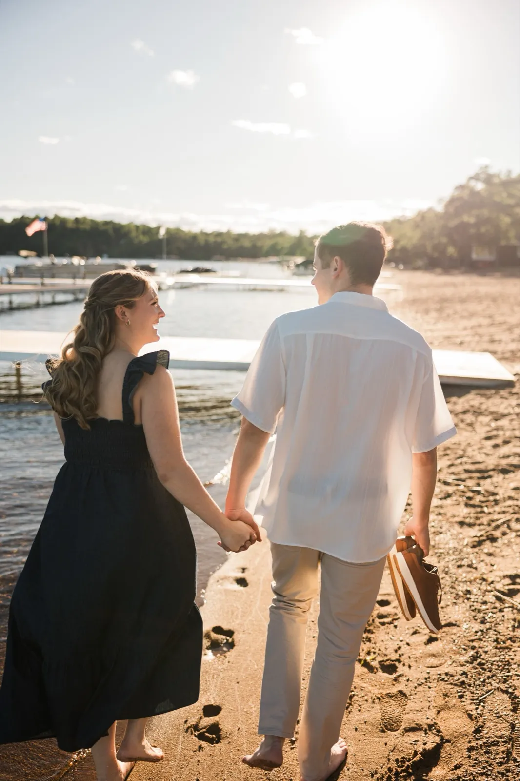 An engaged couple walk away along the sunlit Gull Lake beach holding hands, leaving footprints in the sand — Tim Larsen Photography