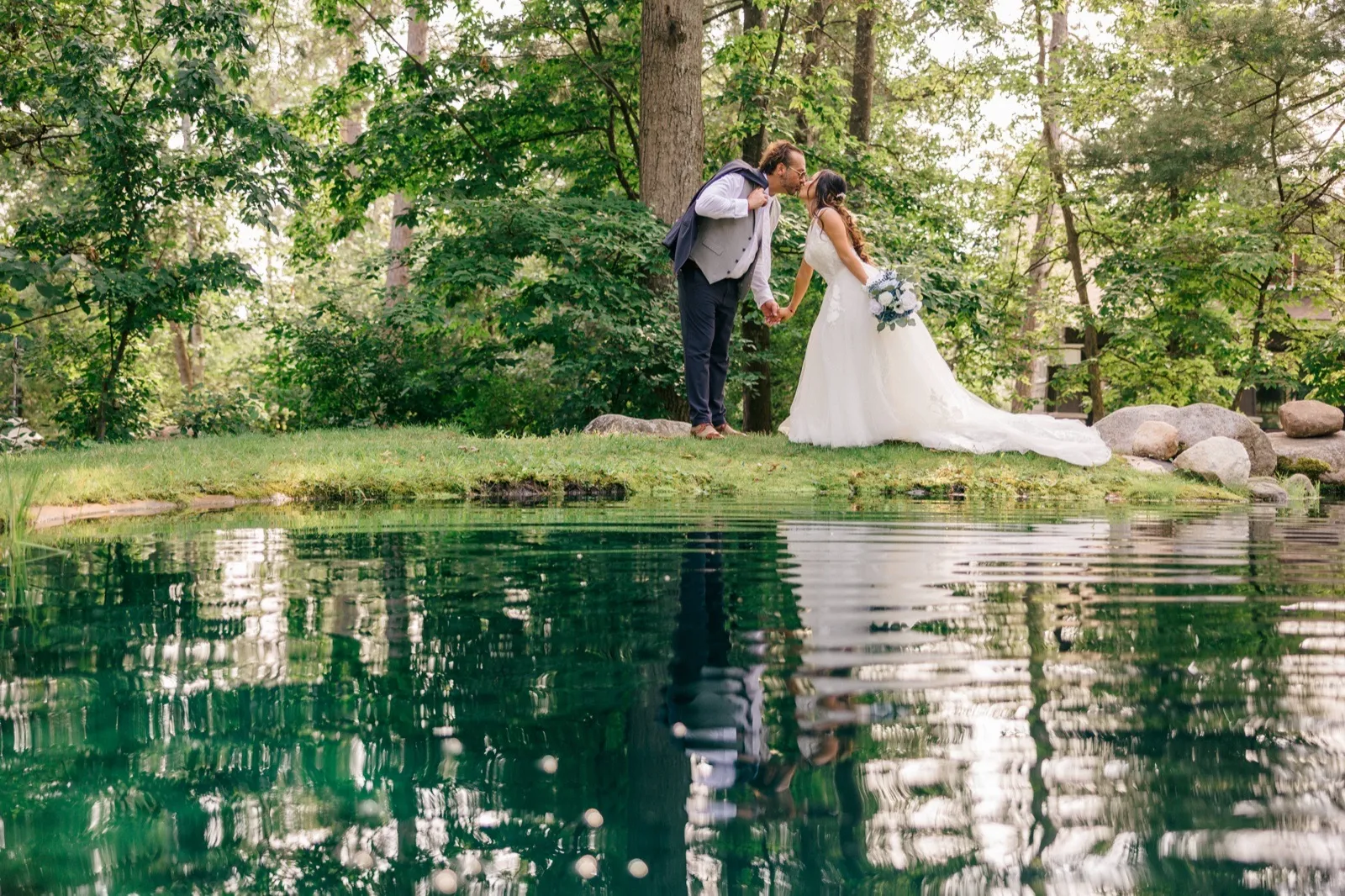 The bride and groom kiss on a grassy bank surrounded by tall trees, their figures and the forest mirrored in the still pond below — Tim Larsen Photography, Brainerd Lakes MN
