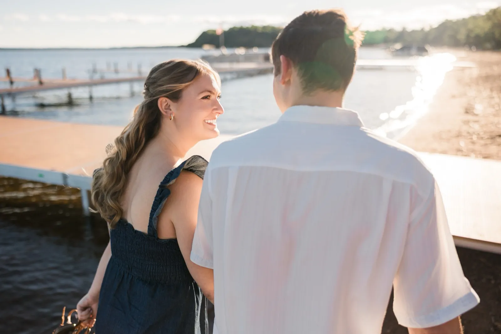 A woman smiles warmly at her fiance, whose back is to the camera, on the Gull Lake shoreline with lens flare — Tim Larsen Photography