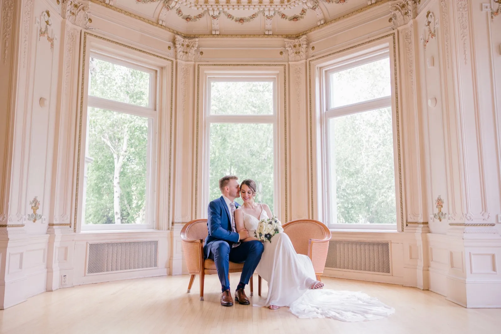 The groom leans in to kiss the bride's temple as they sit together on a small settee in an ornate parlor with tall arched windows and gilded ceiling details at the American Swedish Institute. — Tim Larsen Photography, Minneapolis MN