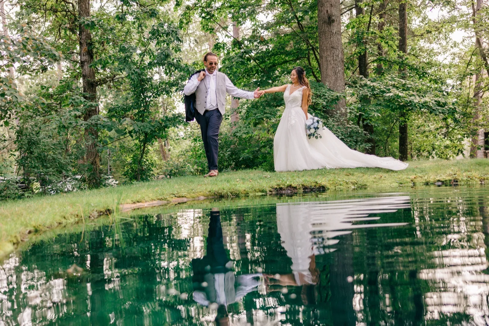 The groom leads the bride by the hand along a grassy bank above a still pond that mirrors them both beneath tall trees at Grand View Lodge — Tim Larsen Photography, Brainerd Lakes MN