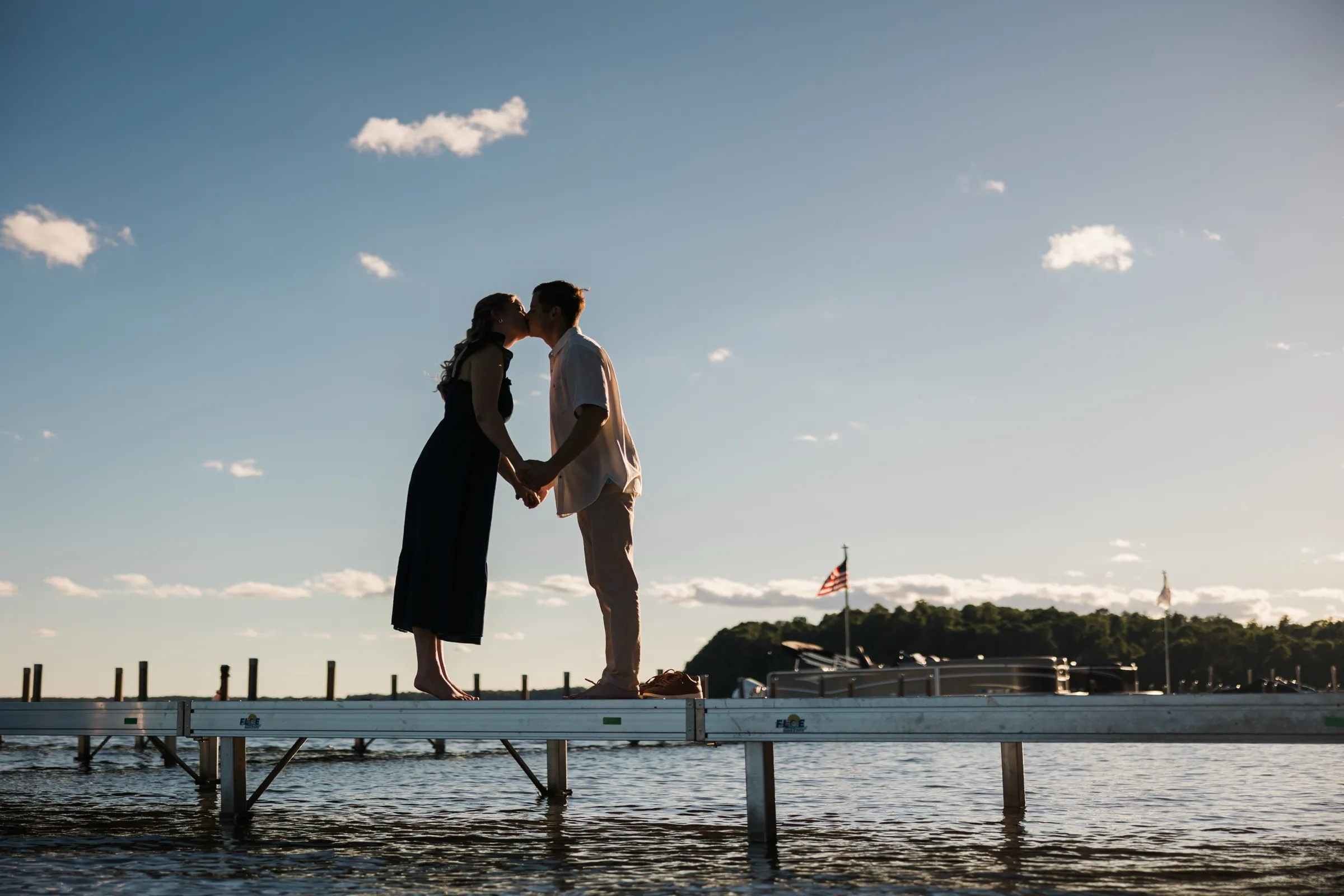 An engaged couple kiss in silhouette on a long dock extending over Gull Lake, against a wide blue sky — Tim Larsen Photography