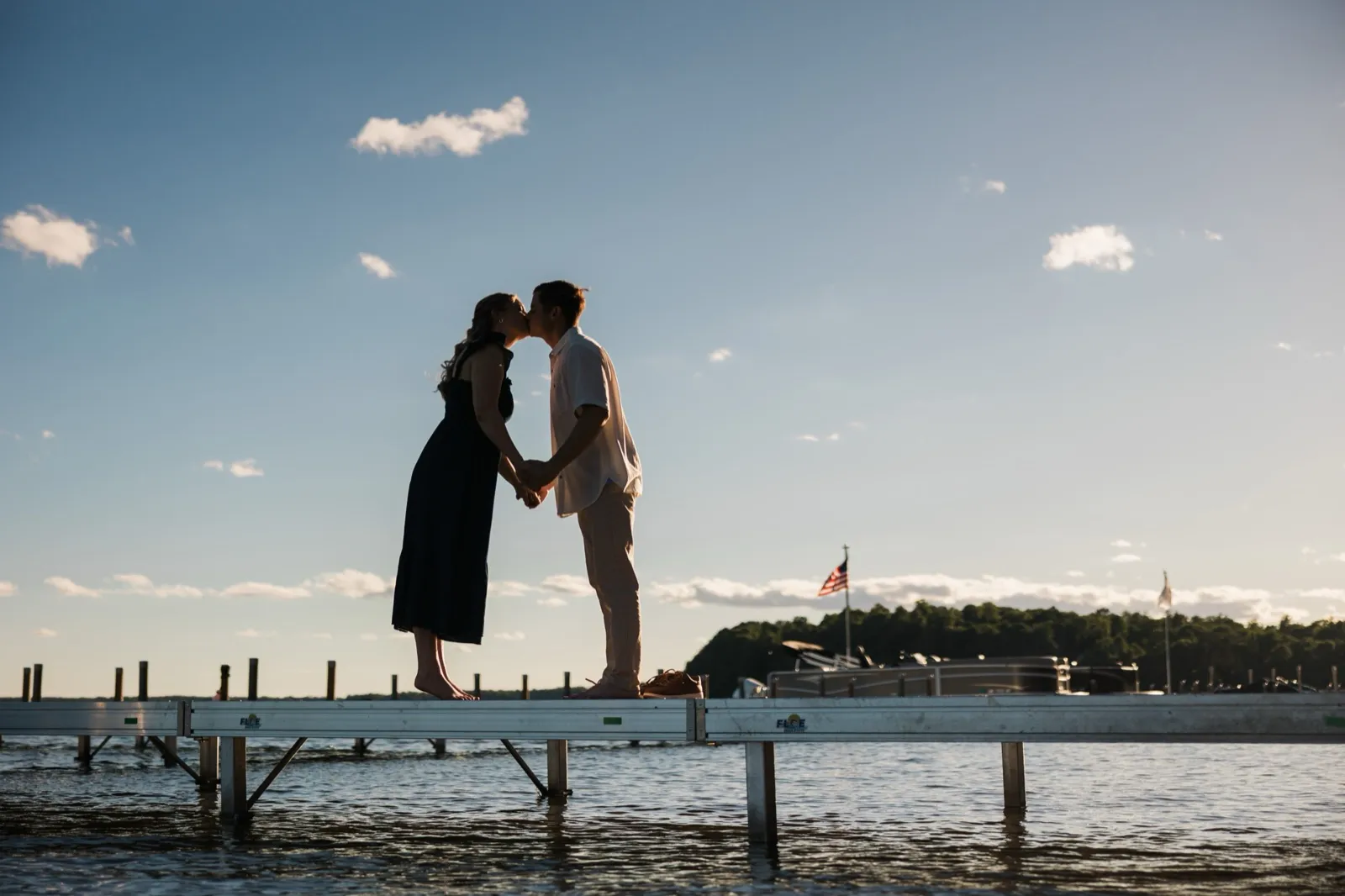 An engaged couple kiss in silhouette on a long dock extending over Gull Lake, against a wide blue sky — Tim Larsen Photography