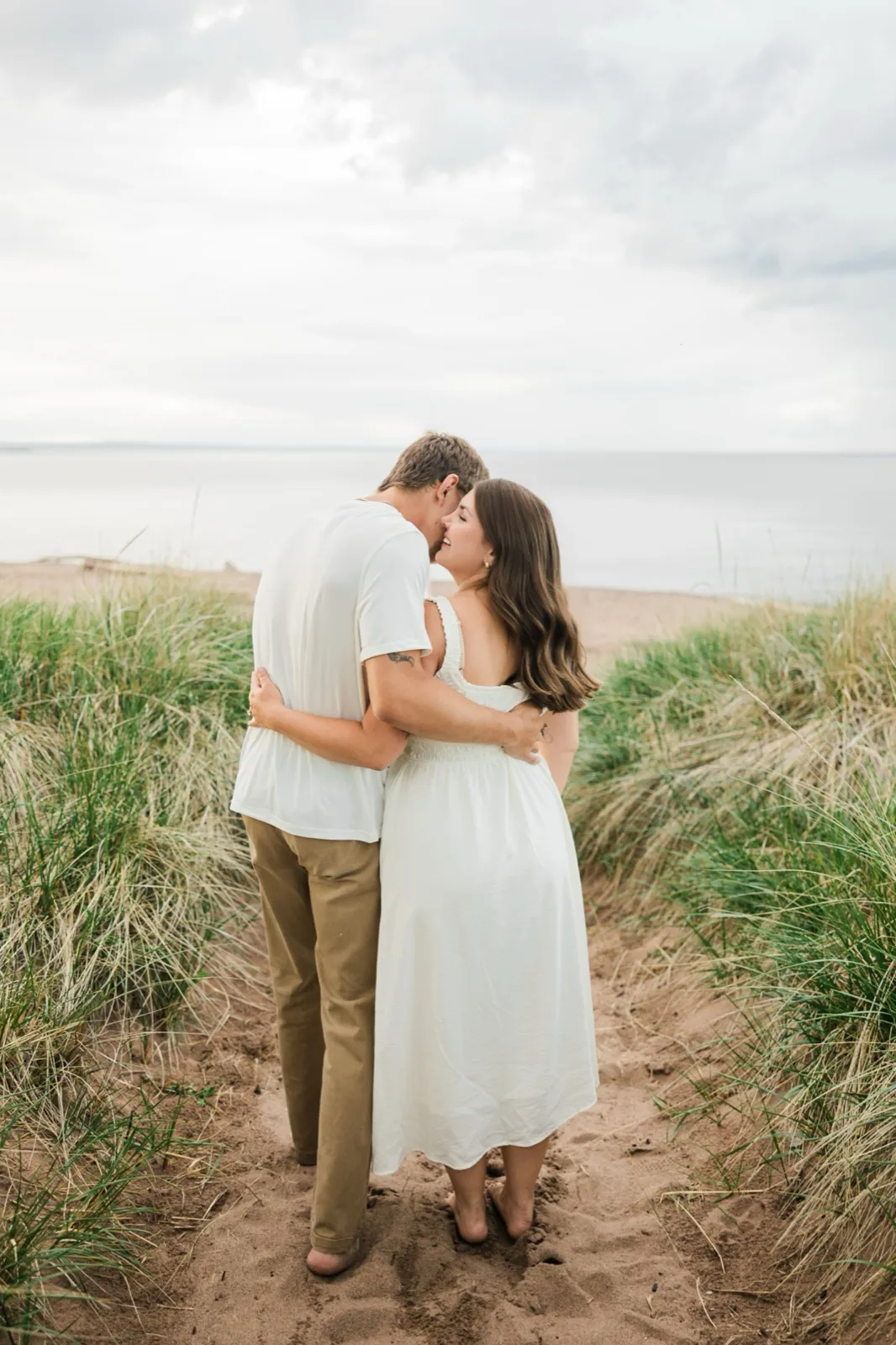 From behind, Reeden and Jacklyn walk the dune-grass path with their arms around each other, turning to kiss as Lake Superior opens ahead — Tim Larsen Photography