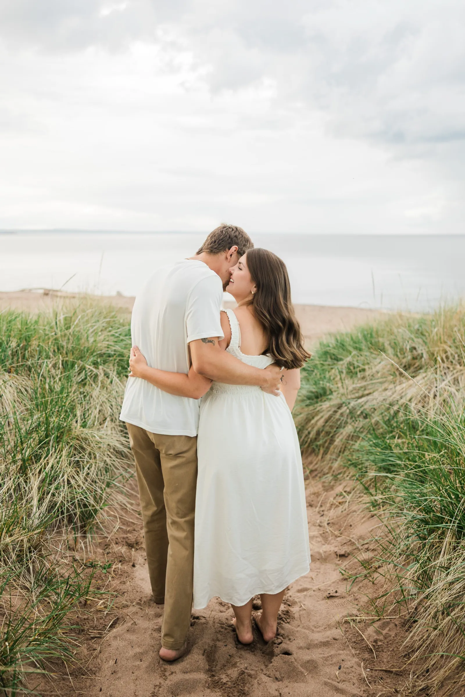 From behind, Reeden and Jacklyn walk the dune-grass path with their arms around each other, turning to kiss as Lake Superior opens ahead — Tim Larsen Photography