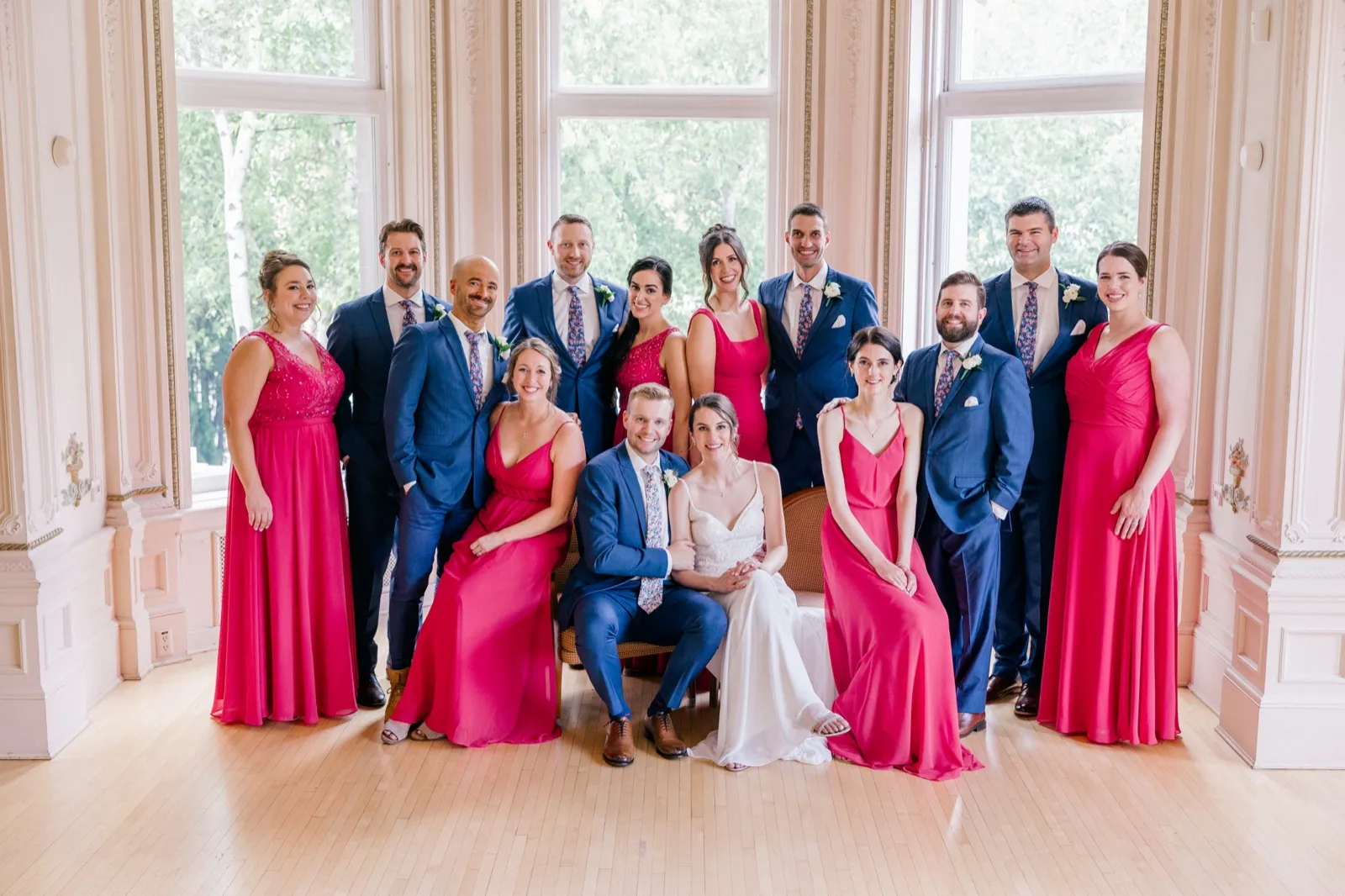 The bridal party of seven women in fuchsia gowns and six men in navy suits gathers around the seated couple in a grand window-lit room with ornate plaster walls. — Tim Larsen Photography, Minneapolis MN