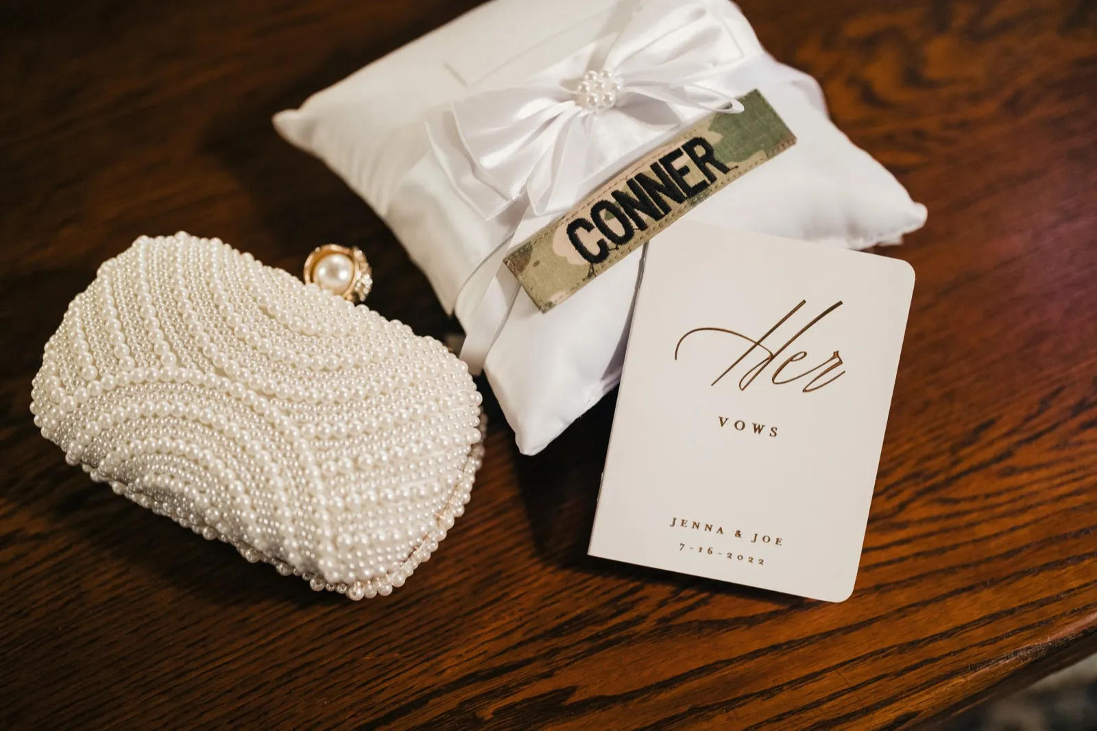 A pearl-beaded clutch, a white satin ring pillow with a military name tape reading 'CONNER,' and a vow book labeled 'Her Vows — Jenna & Joe, 7-16-2022' arranged on a dark wood surface. — Tim Larsen Photography, Brainerd Lakes MN