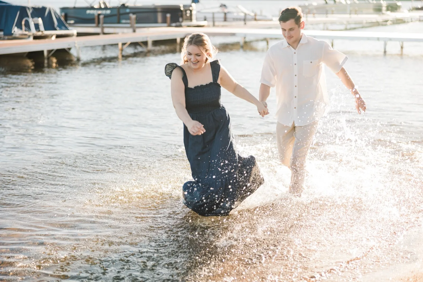 A laughing engaged couple run hand in hand through the Gull Lake shallows, kicking up spray, backlit by the sun — Tim Larsen Photography