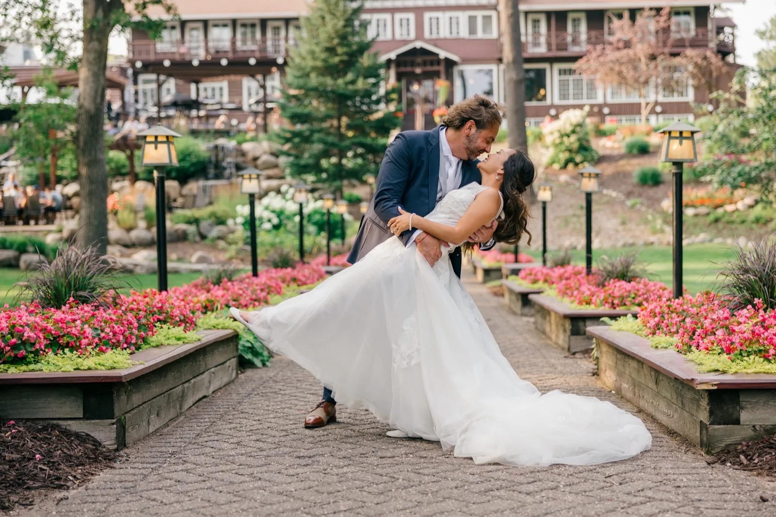 The groom dips the bride backward as she laughs on a stone garden path lined with pink flowers and lantern posts, the timbered Grand View Lodge rising behind them — Tim Larsen Photography, Brainerd Lakes MN