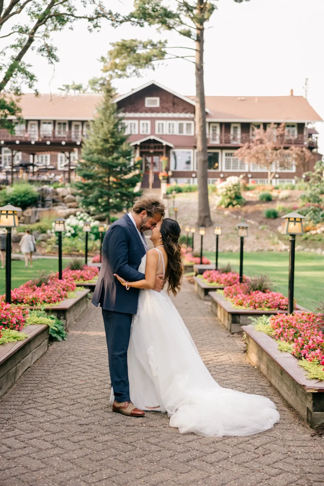 The bride and groom stand forehead-to-forehead on a symmetrical brick pathway lined with lanterns and pink floral beds, with the historic Grand View Lodge rising behind them — Tim Larsen Photography, Brainerd Lakes MN