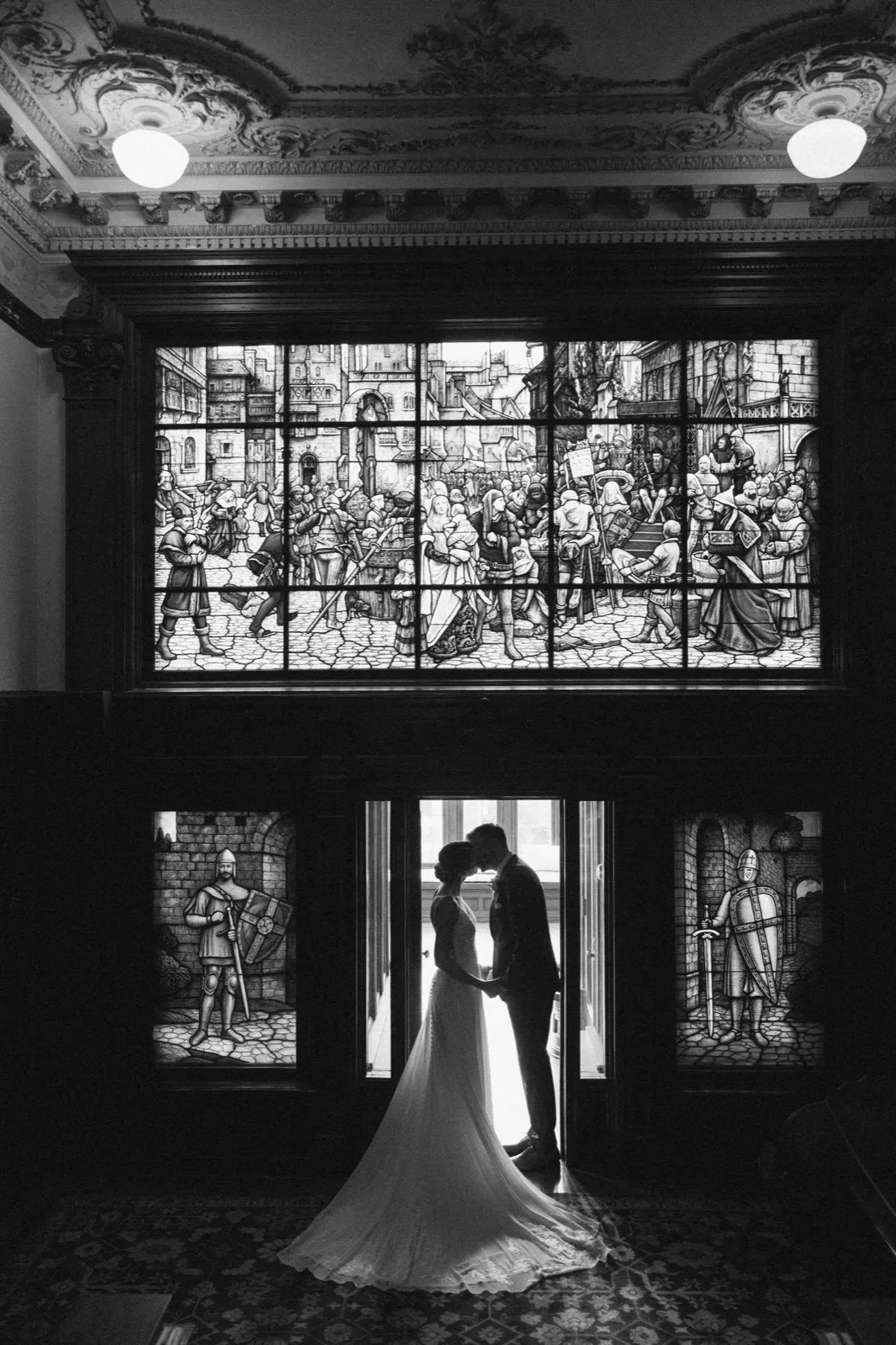 The bride and groom stand silhouetted in a doorway beneath a vast stained glass window depicting a medieval scene, framed by ornate ceiling plasterwork at the American Swedish Institute. — Tim Larsen Photography, Minneapolis MN