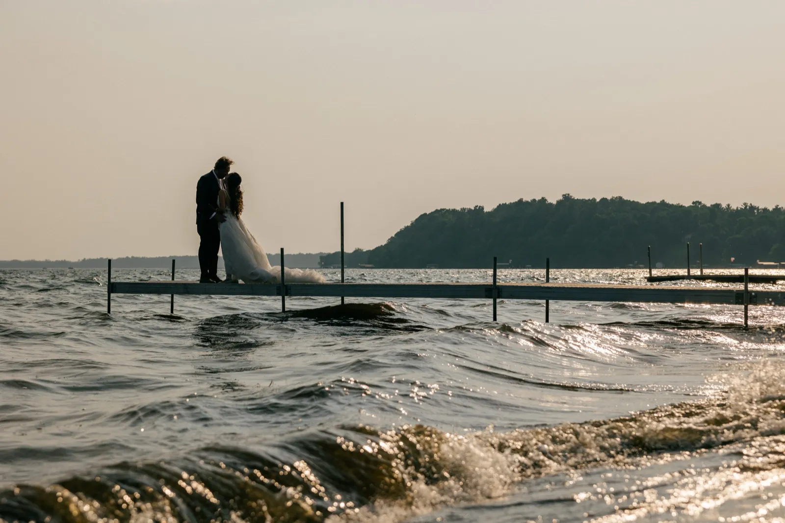 The bride and groom stand close together on the long Grand View Lodge dock at Gull Lake at dusk, their silhouettes set against a hazy sky as waves break in the foreground — Tim Larsen Photography, Brainerd Lakes MN