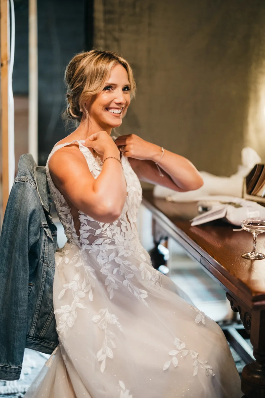 The bride laughs as she fastens a necklace behind her neck while seated at a wooden vanity, her jean jacket draped over the chair and a coupe glass on the table beside her. — Tim Larsen Photography, Brainerd Lakes MN