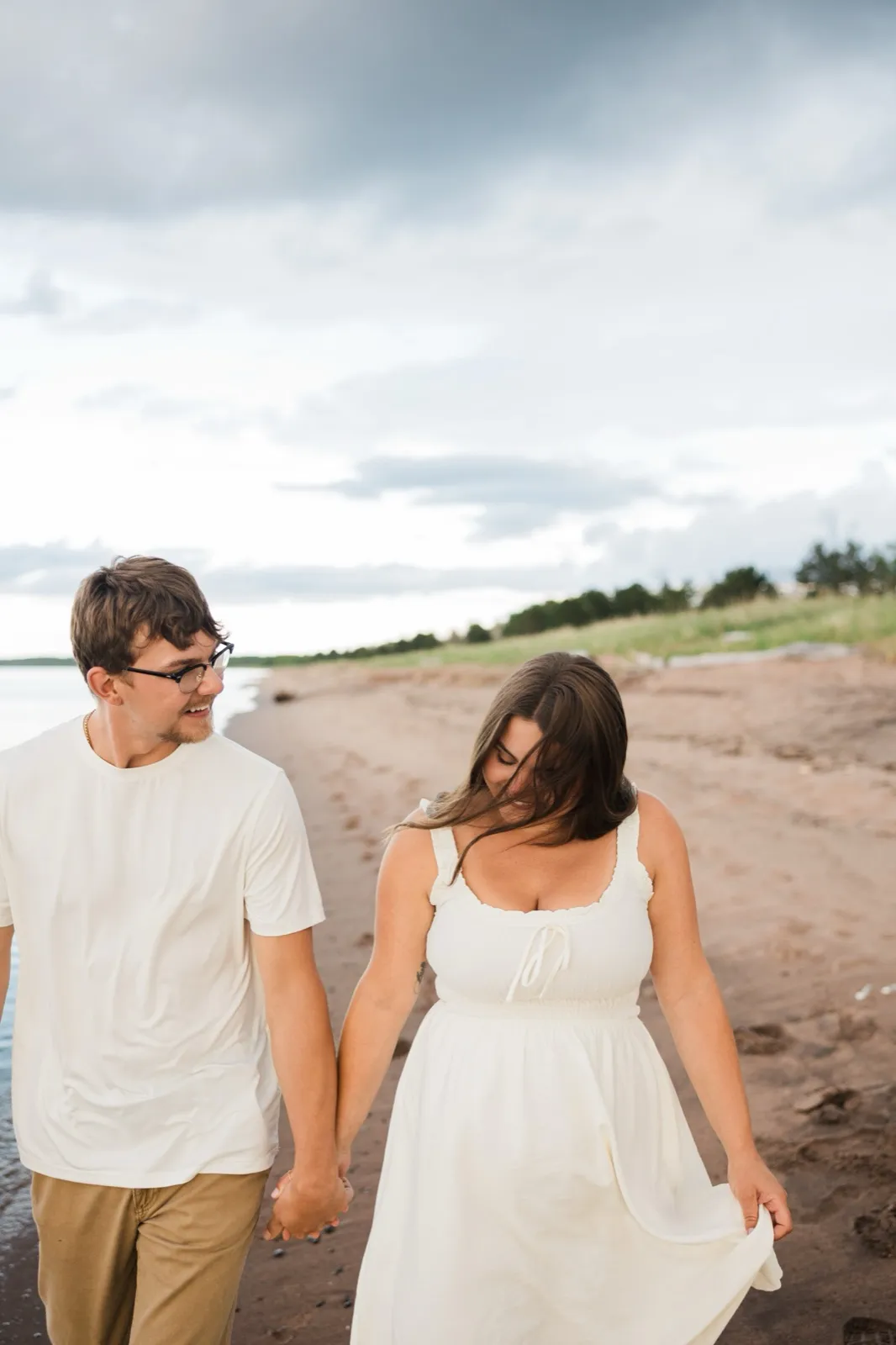 Reeden and Jacklyn walk the beach hand in hand, Jacklyn's hair blown across her face as he watches her — Tim Larsen Photography