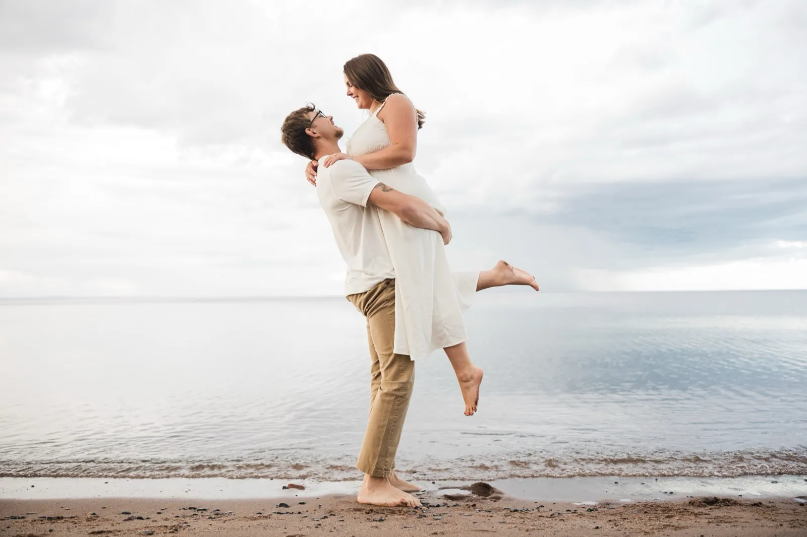 Reeden lifts Jacklyn at the Lake Superior waterline as they gaze at each other, the calm lake silver behind them — Tim Larsen Photography