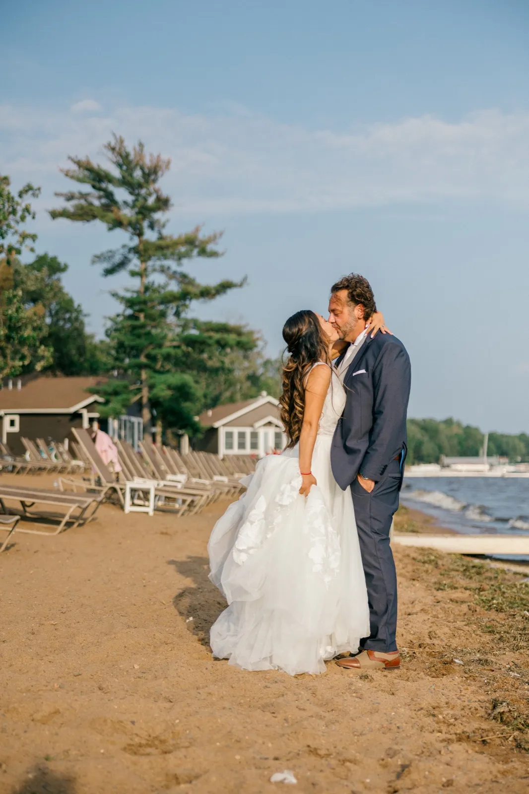 The bride and groom share a kiss on the sandy beach at Grand View Lodge with lounge chairs, lakeside cabins, and Gull Lake visible behind them — Tim Larsen Photography, Brainerd Lakes MN