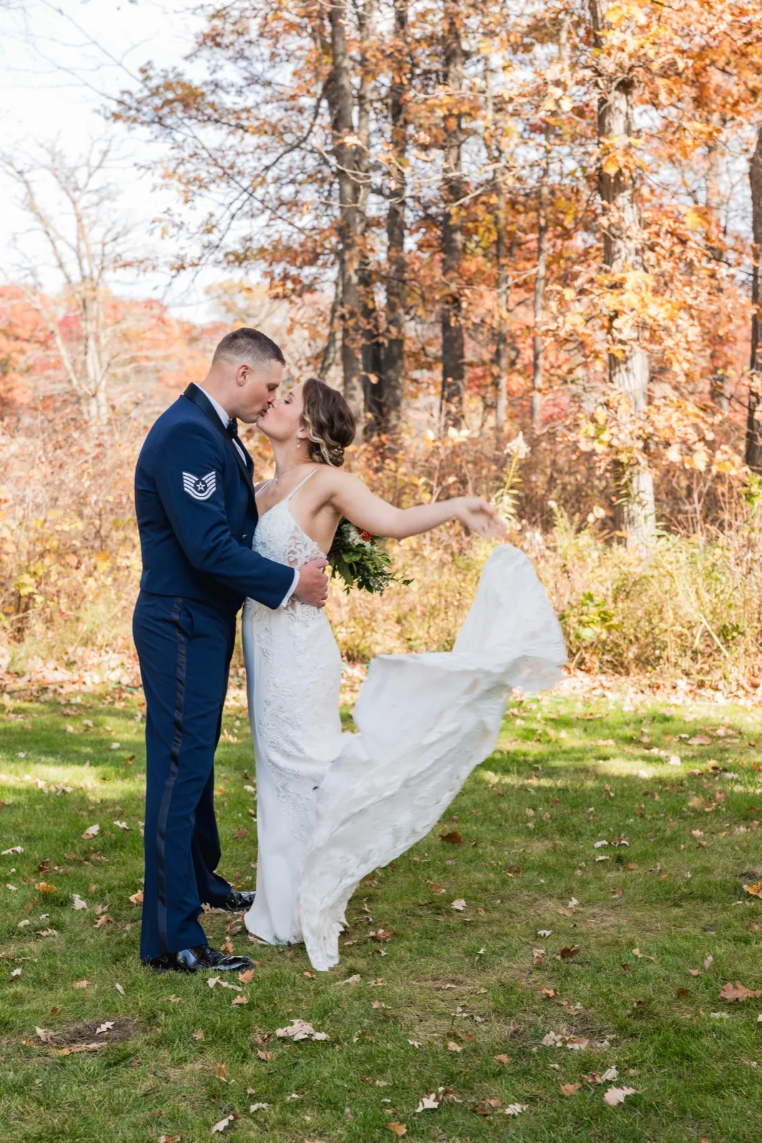 The bride kisses the groom while sweeping her gown out to one side against a backdrop of autumn trees — Tim Larsen Photography, Brainerd Lakes MN
