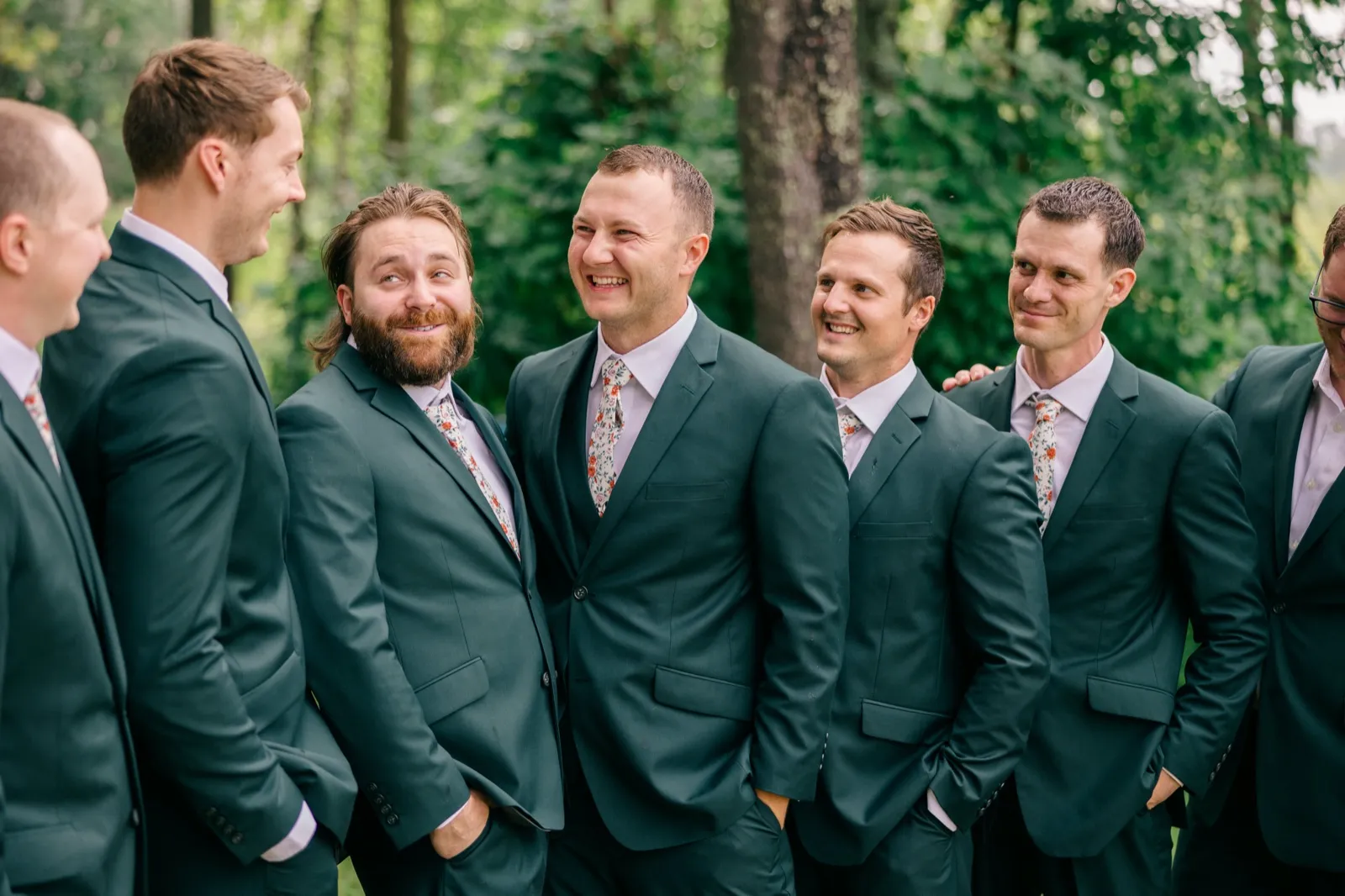 Eric laughs broadly while his groomsmen, dressed in matching green suits with floral ties, share the joke beside him in a wooded clearing — Tim Larsen Photography, Brainerd Lakes MN