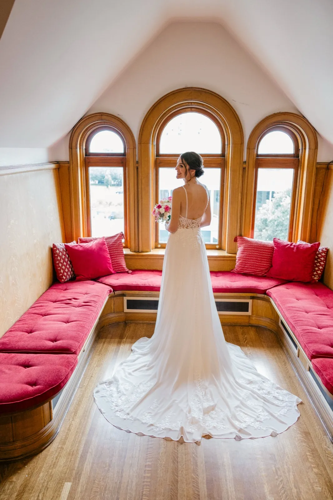 The bride stands centered in a symmetrical turret alcove with three arched windows, glancing over her bare back as her lace train fans across the hardwood floor. — Tim Larsen Photography, Minneapolis MN