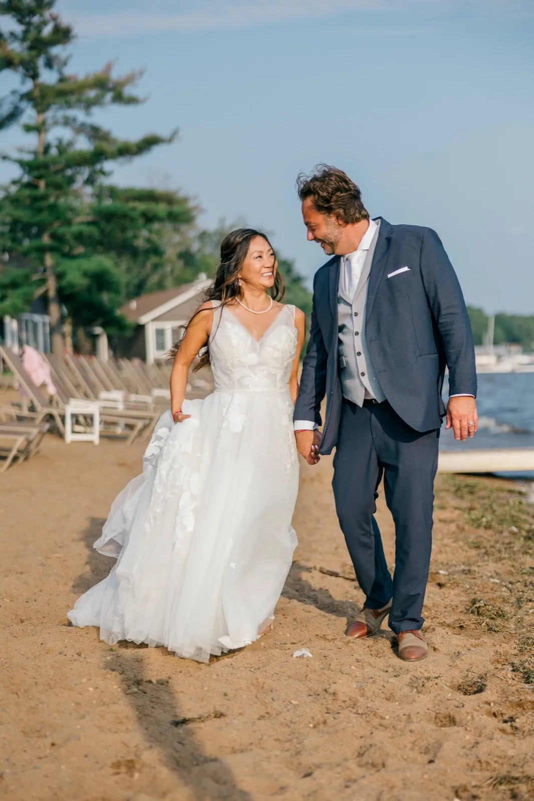 The bride and groom walk hand in hand along a sandy lakeshore at Grand View Lodge, smiling at each other as her tulle gown trails through the sand — Tim Larsen Photography, Brainerd Lakes MN
