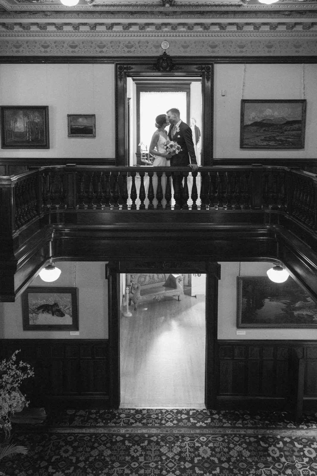 A bride and groom kiss on an ornate second-floor balcony of a historic mansion, framed symmetrically by carved wood railings, doorways, and oil paintings on either side. — Tim Larsen Photography, Minneapolis MN