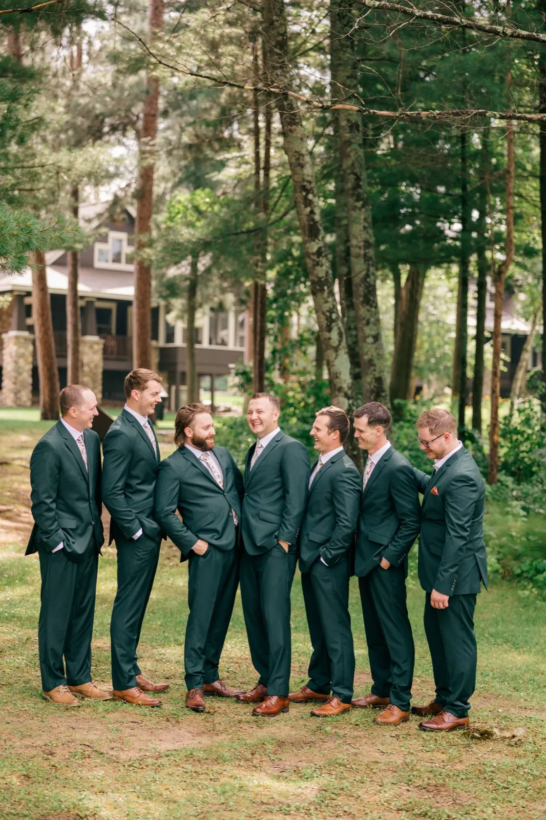 The groom and his seven groomsmen in matching green suits laugh together on a grassy lawn surrounded by tall pines, with Grand View Lodge visible in the background