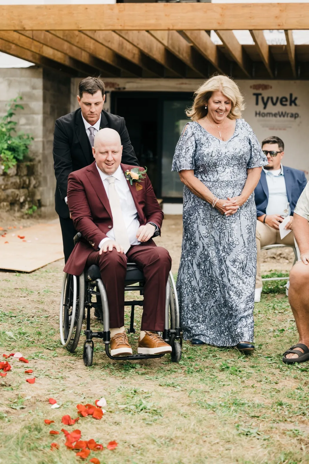 The groom, seated in a wheelchair and wearing a burgundy suit, is escorted down a petal-strewn aisle by a man in a black suit while his mother walks beside him smiling. — Tim Larsen Photography, Brainerd Lakes MN
