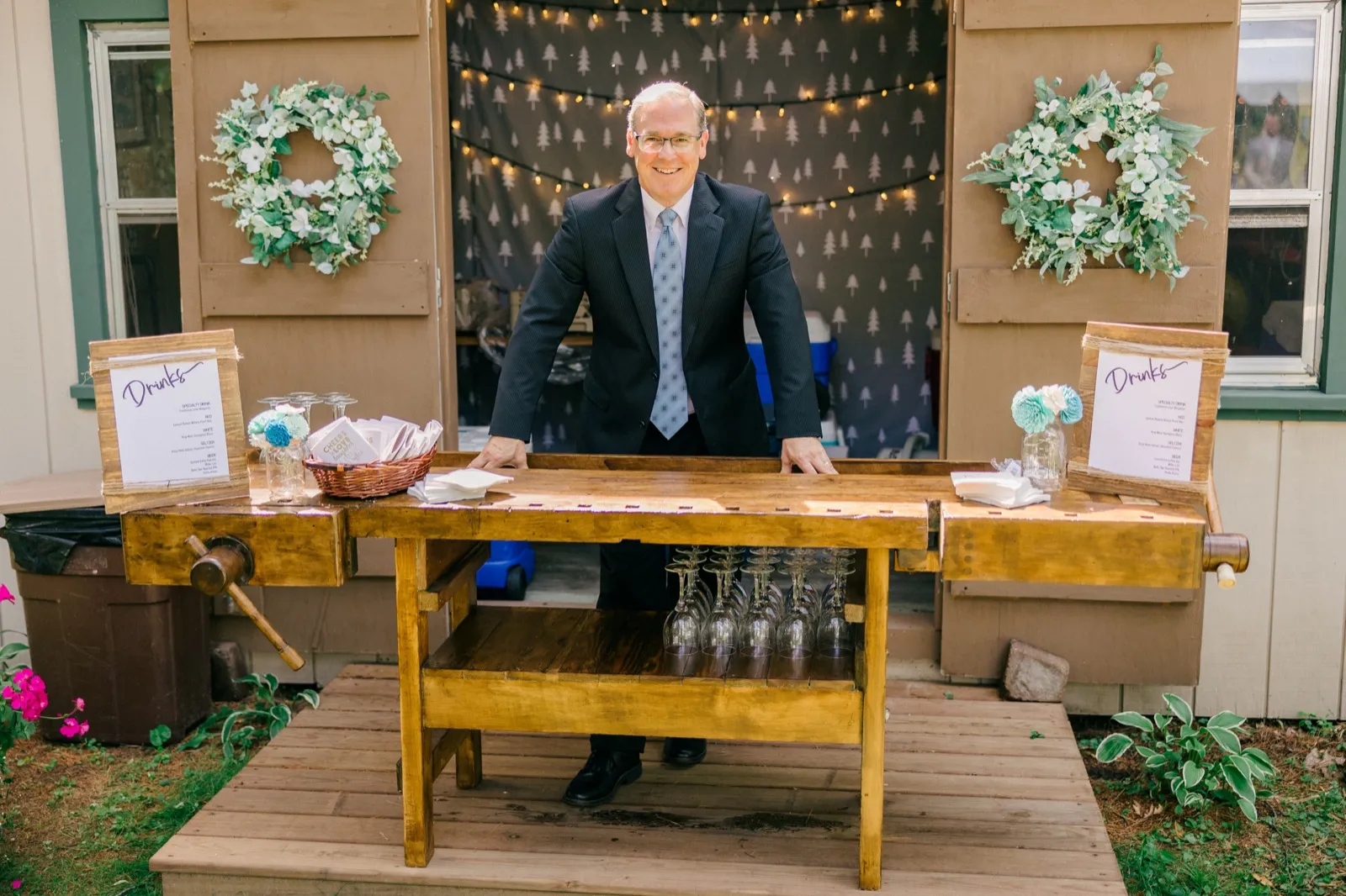 The wooden bar the bride's father built for the wedding, set with wine glasses and drink menus beneath floral wreaths and string lights, a man in a dark suit and tie smiling behind it. — Tim Larsen Photography, Brainerd Lakes MN