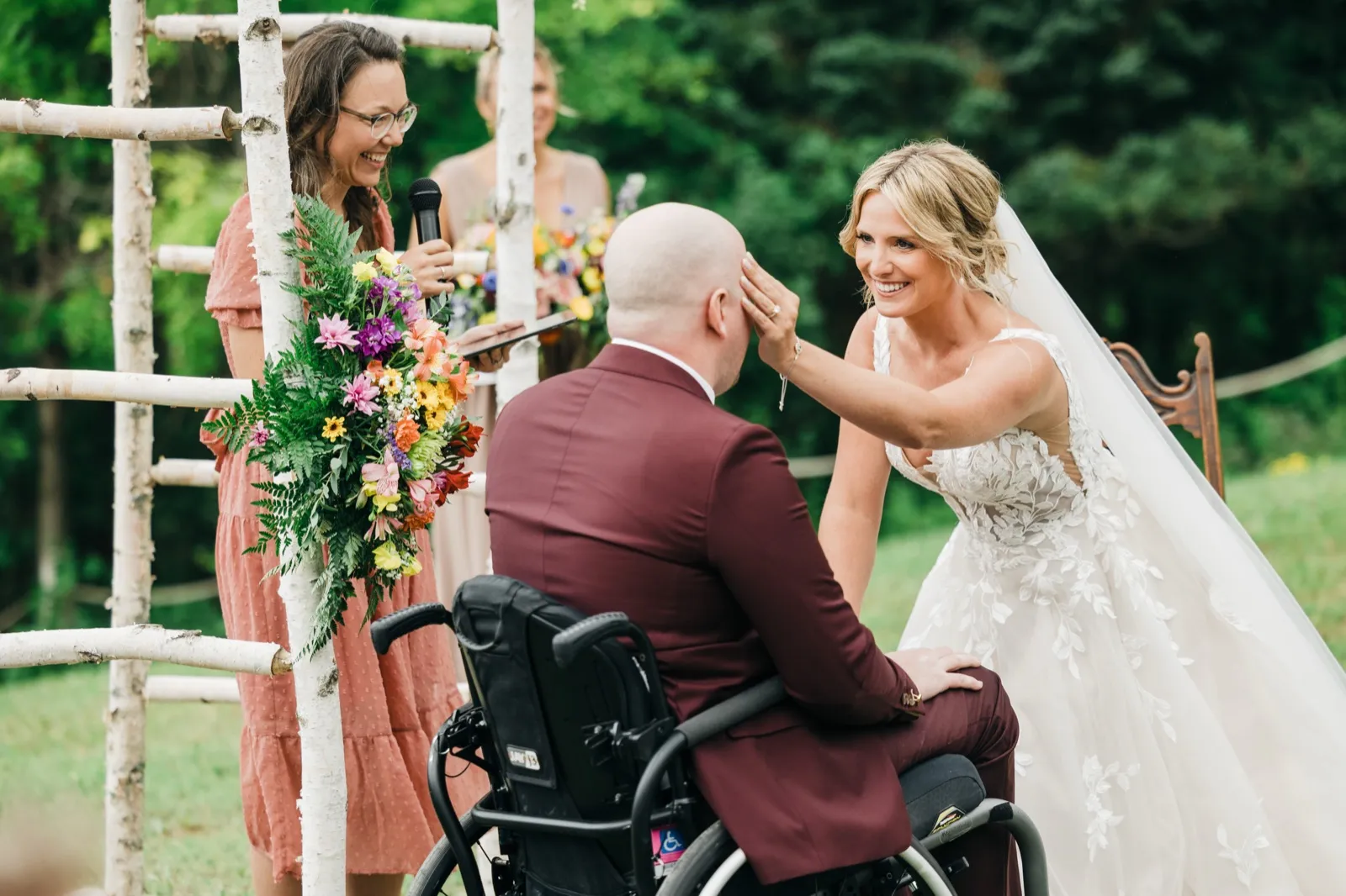 The bride leans forward from her chair to gently wipe the groom's brow as he sits in his wheelchair during their outdoor ceremony, while the officiant laughs nearby holding a microphone. — Tim Larsen Photography, Brainerd Lakes MN