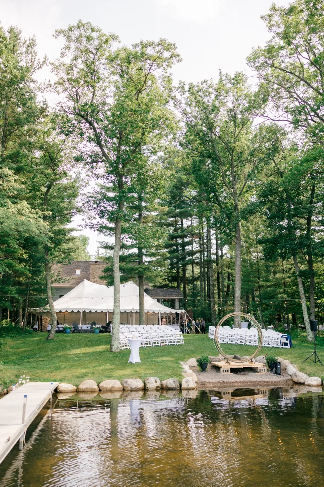 A wide view across a calm lake shows a sailcloth tent, a circular wooden ceremony arch, and rows of white chairs set among tall pines on a private cabin property. — Tim Larsen Photography, Brainerd Lakes MN