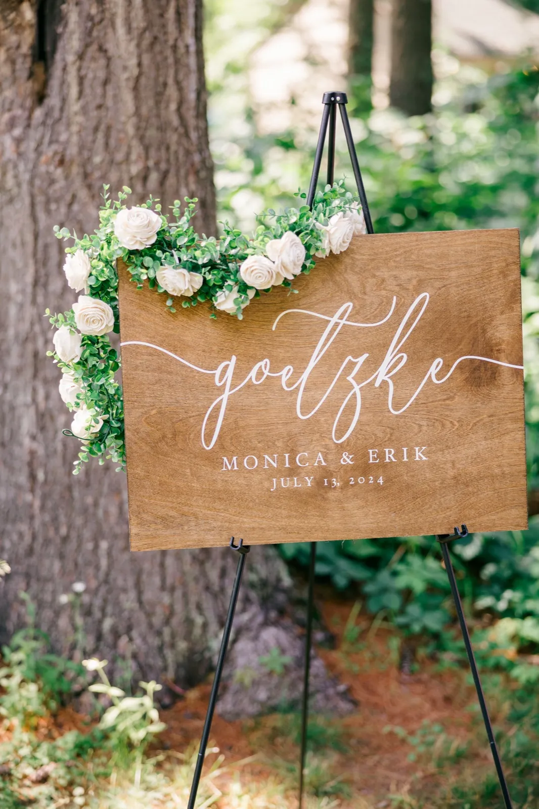 A wooden welcome sign reading 'Goetzke, Monica & Erik, July 13, 2024' rests on a black easel, draped with eucalyptus and ivory roses, set against a large tree trunk in dappled forest light. — Tim Larsen Photography, Brainerd Lakes MN