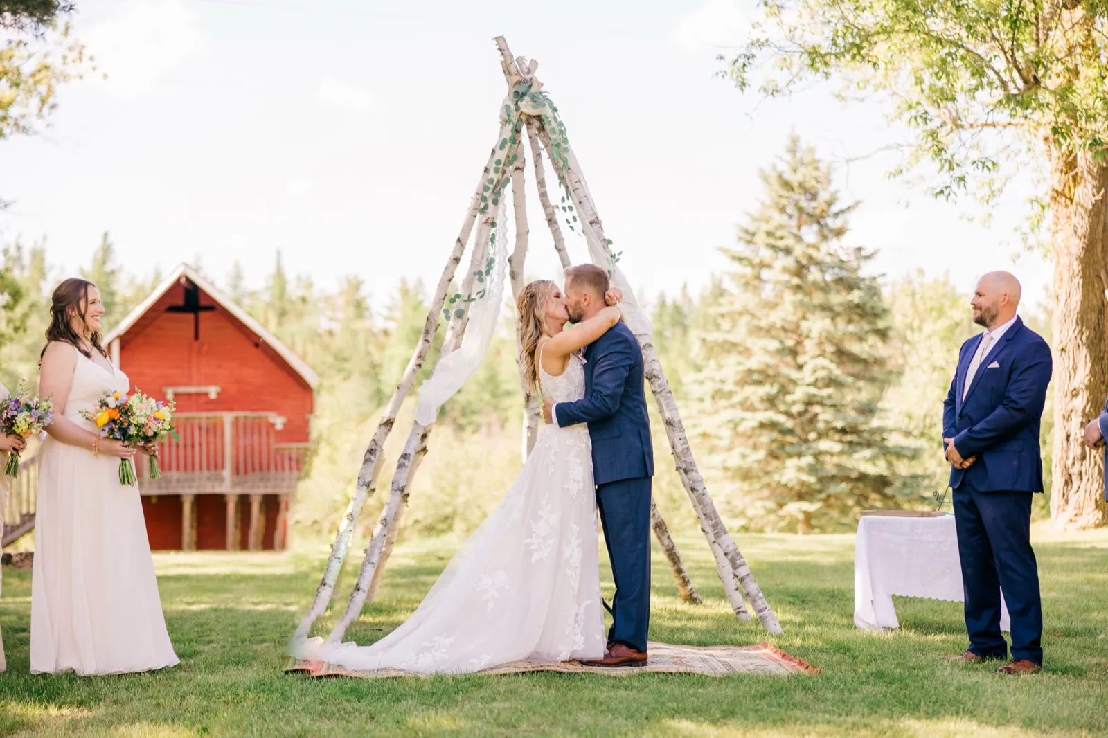 The bride and groom share their first kiss beneath a birch-pole teepee arch on a sunlit lawn, with a bridesmaid holding a wildflower bouquet on the left and a red barn visible in the background — Tim Larsen Photography, Brainerd Lakes MN
