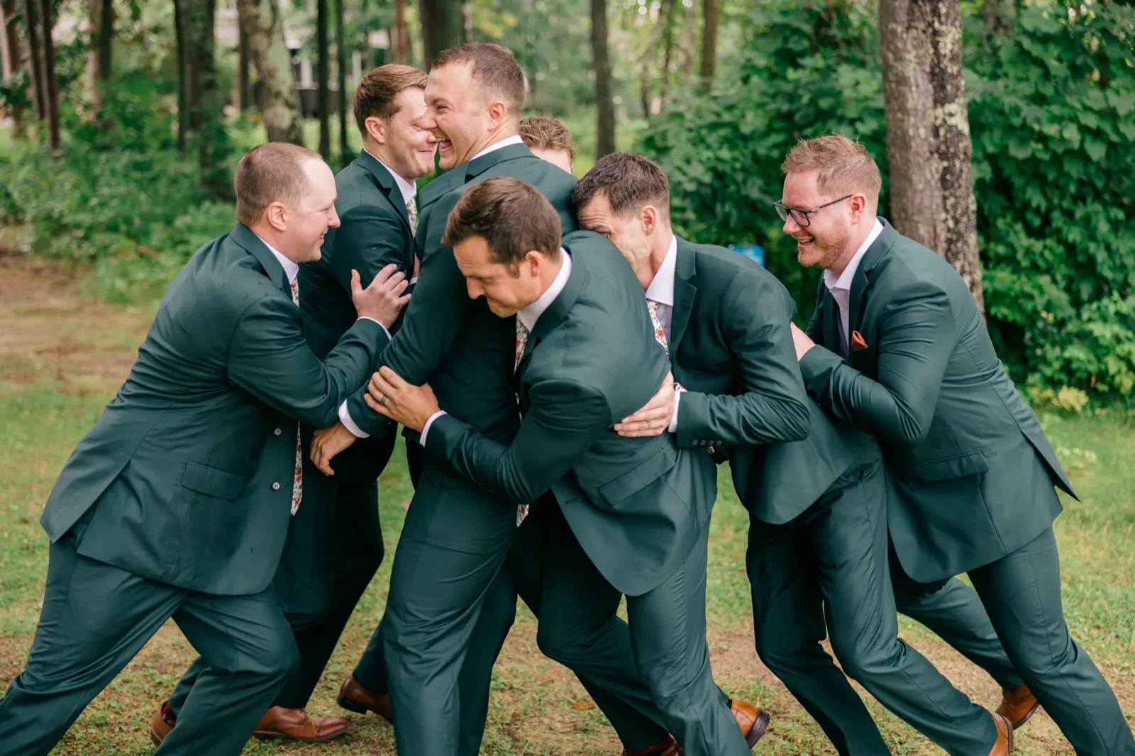 Groomsmen in matching green suits pile onto Eric in a laughing scrum on the grass at the edge of a wooded clearing — Tim Larsen Photography, Brainerd Lakes MN