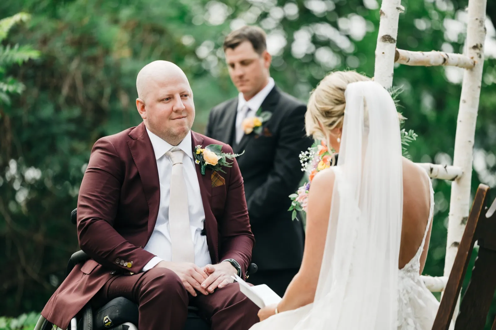 The groom, seated in a wheelchair and wearing a burgundy suit, looks toward his bride as she reads her vows during an outdoor ceremony, with the best man and a birch arch behind him. — Tim Larsen Photography, Brainerd Lakes MN