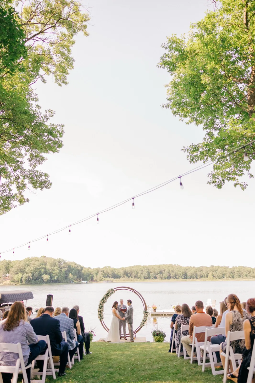 A bride and groom stand at a circular floral arch exchanging vows beside a lake, framed by tall trees and seated guests on a sunny afternoon. — Tim Larsen Photography, Brainerd Lakes MN