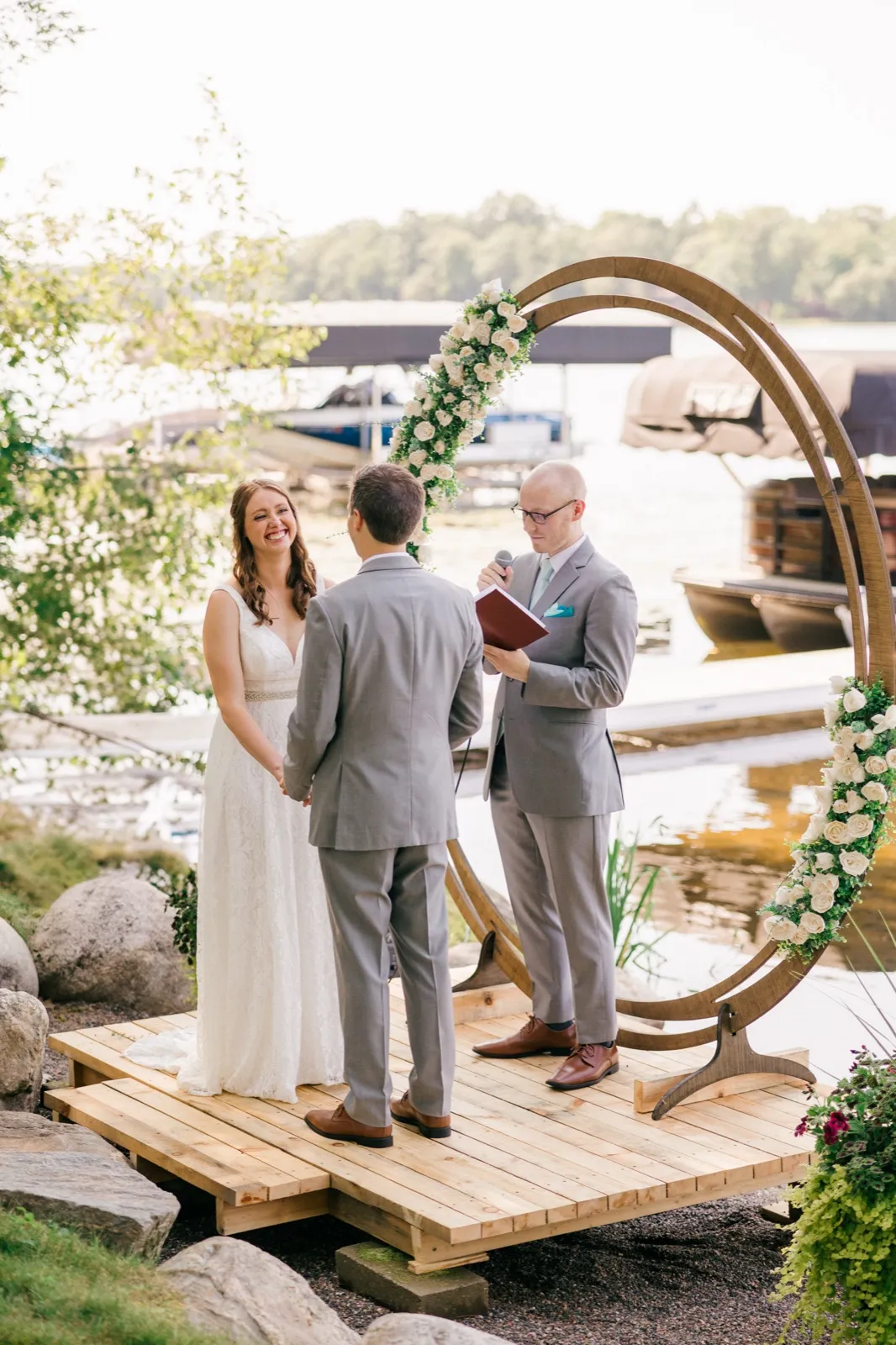The bride laughs brightly while holding the groom's hand as the officiant reads from a book beneath a circular floral arch on a lakeside platform. — Tim Larsen Photography, Brainerd Lakes MN