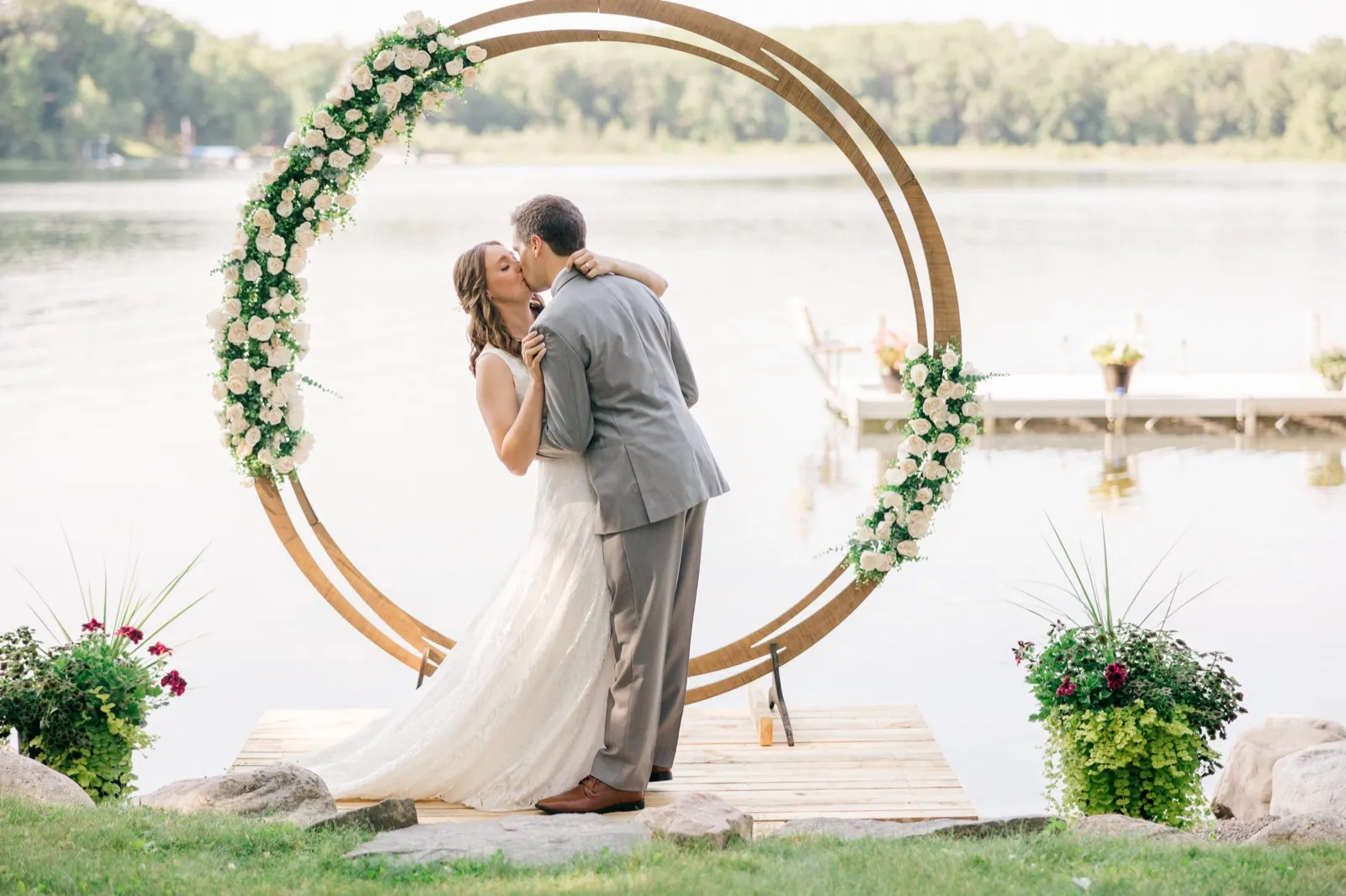 The bride and groom share their first kiss inside a circular floral arch on a wooden platform at the edge of a Brainerd Lakes private cabin shoreline. — Tim Larsen Photography, Brainerd Lakes MN