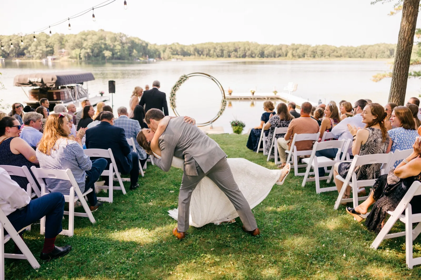 The groom dips the bride into a kiss in the grass aisle while seated guests laugh and applaud, a circular floral arch and lake visible behind them. — Tim Larsen Photography, Brainerd Lakes MN