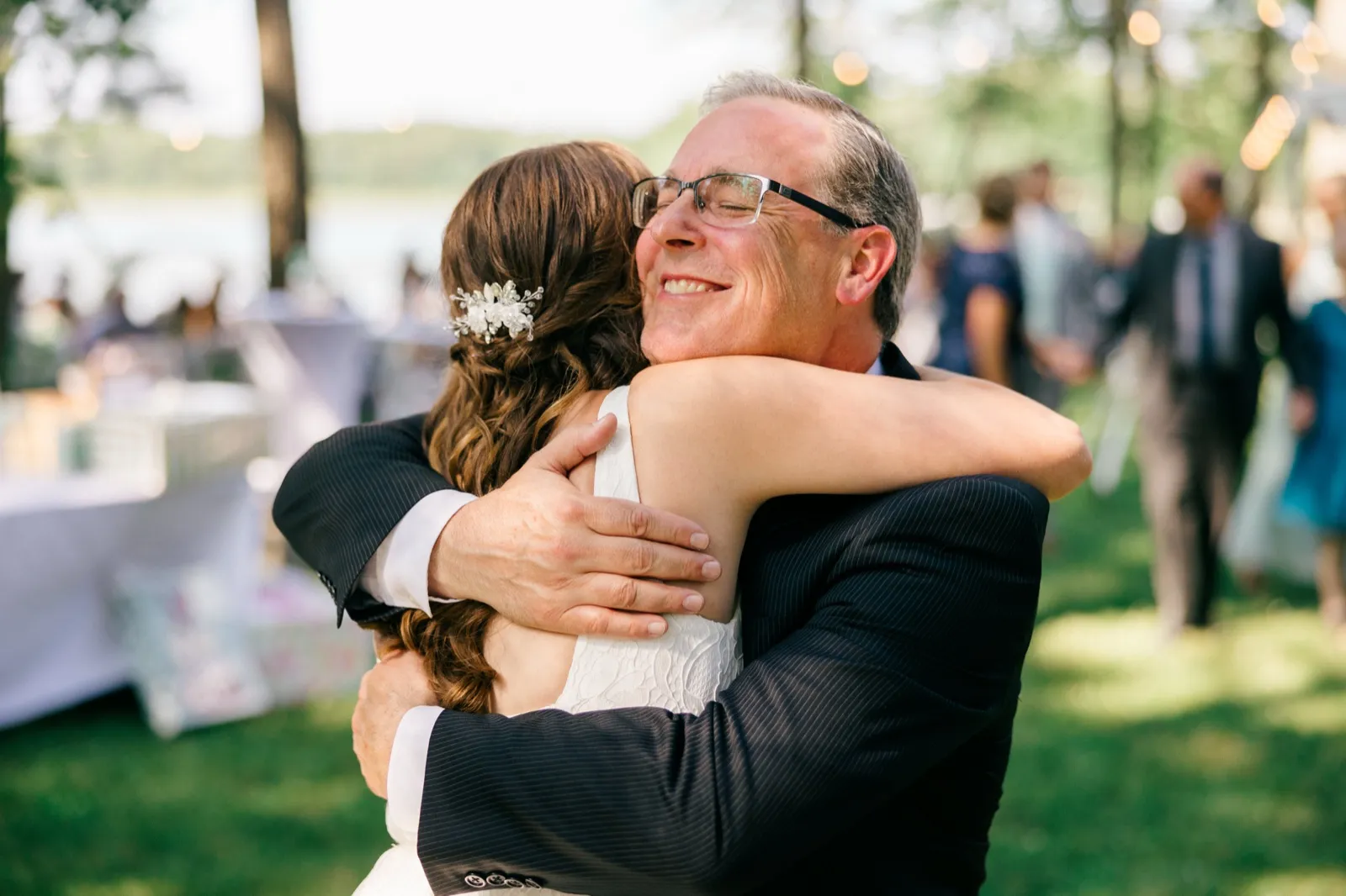 The bride's father holds her in a tight embrace, eyes closed and smiling, on a sunlit lawn with the lake and guests blurred behind them. — Tim Larsen Photography, Brainerd Lakes MN