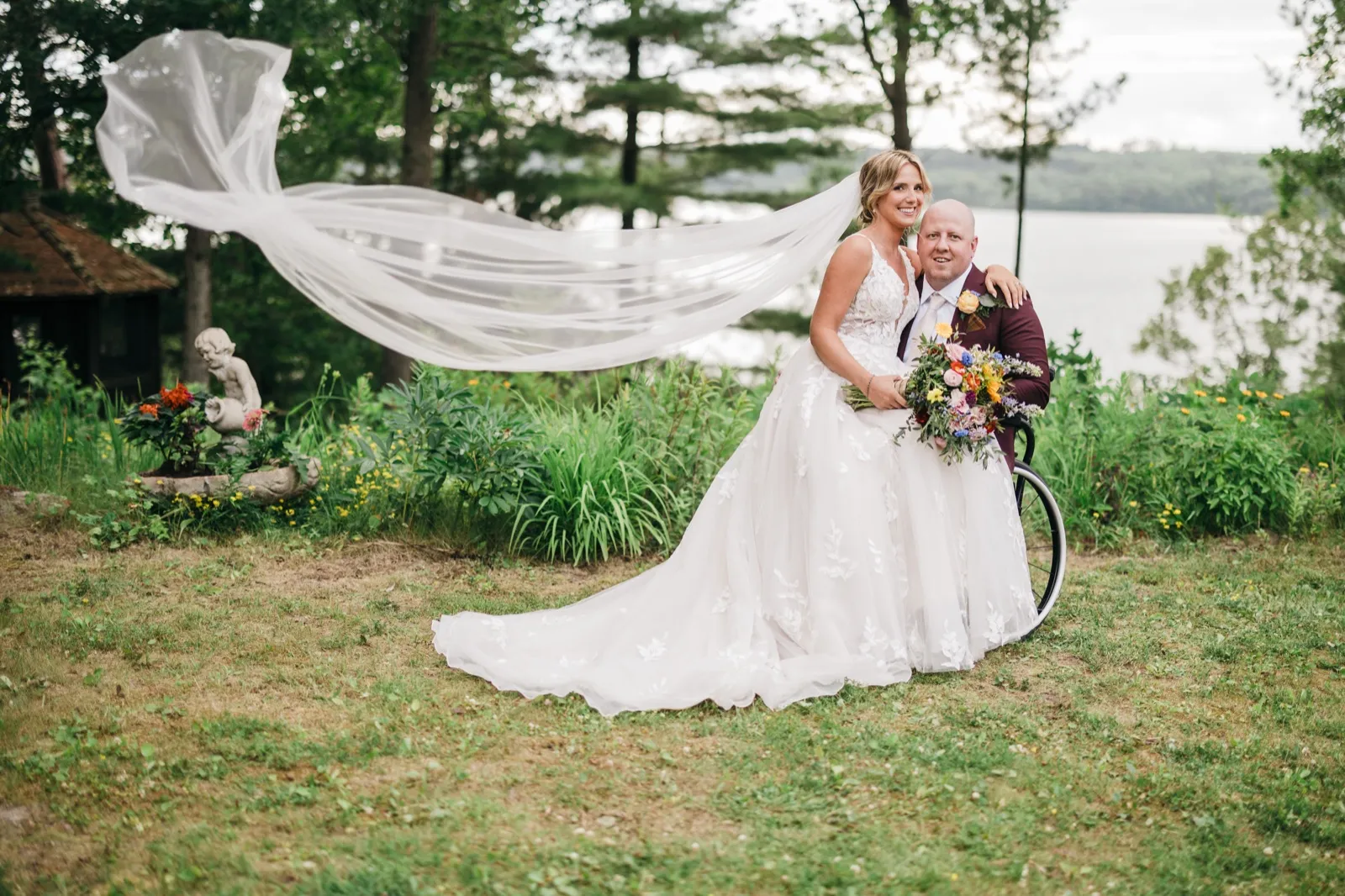 The bride's long veil catches the wind and sweeps across the frame as she stands beside her groom in his wheelchair, holding a colorful bouquet on a lakeside lawn. — Tim Larsen Photography, Brainerd Lakes MN