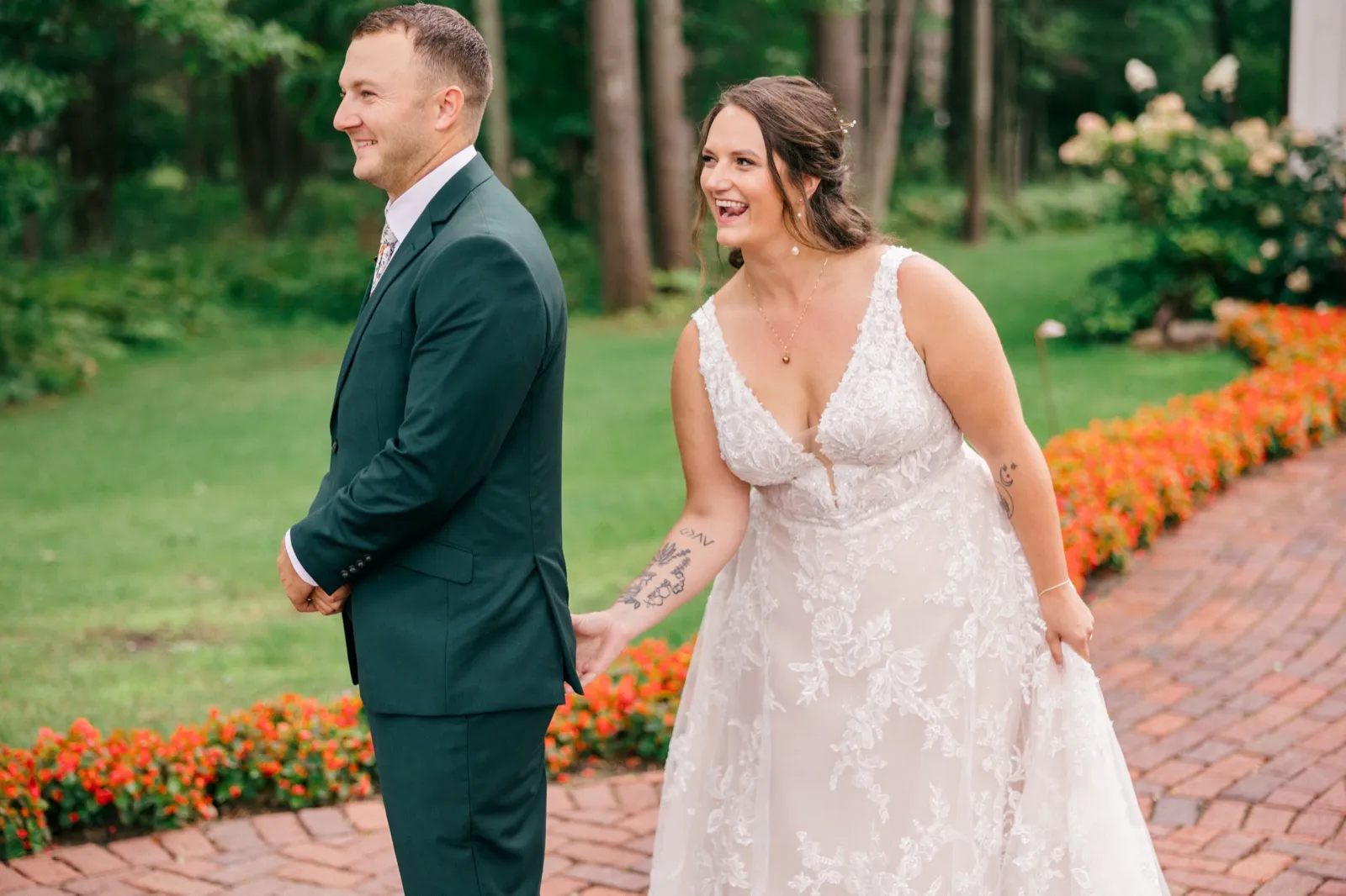 Chelsea laughs openly while standing back-to-back with Eric on a brick garden path lined with red flowers before their first look — Tim Larsen Photography, Brainerd Lakes MN