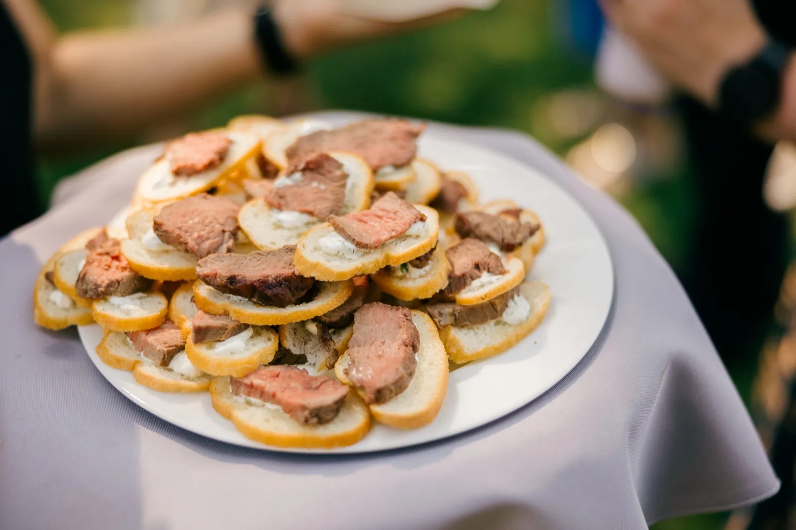 A server holds a white platter piled with beef crostini on sliced baguette during an outdoor cocktail hour. — Tim Larsen Photography, Brainerd Lakes MN