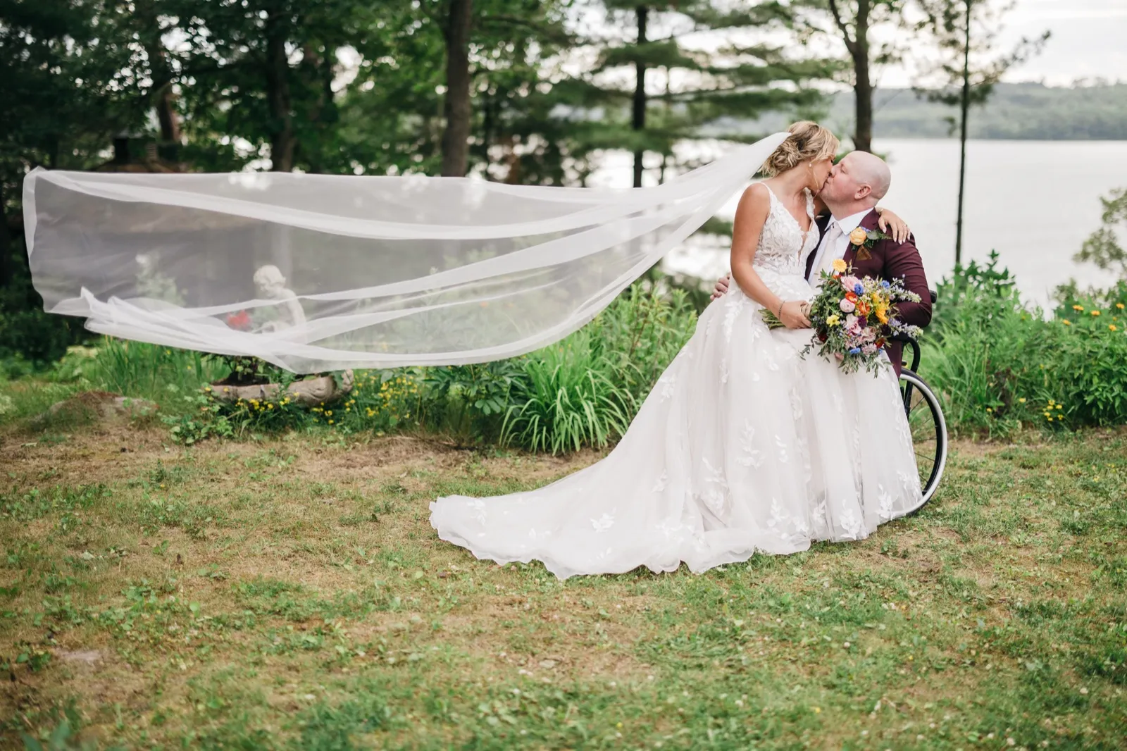 The bride and groom kiss on a lakeside lawn as her long veil catches the wind and stretches dramatically across the frame, with a wheelchair and colorful bouquet beside them. — Tim Larsen Photography, Brainerd Lakes MN