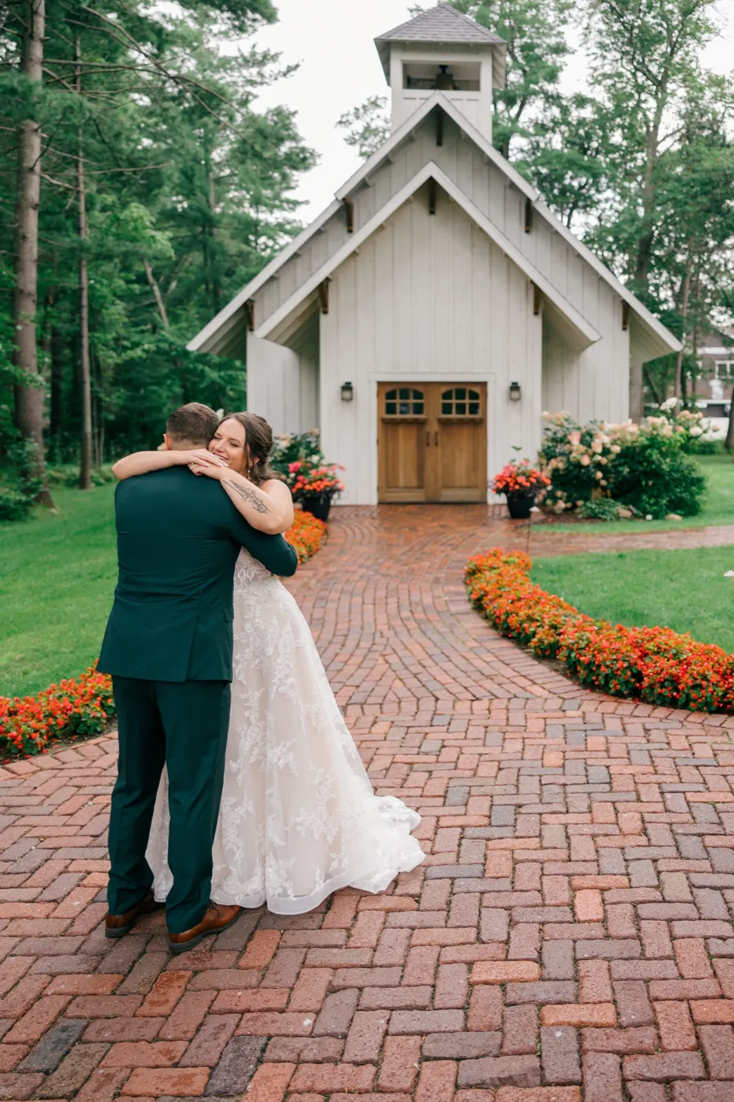 Chelsea smiles with closed eyes as she embraces Eric on a brick walkway leading to Grand View Lodge's white chapel, flanked by red and orange flower beds — Tim Larsen Photography, Brainerd Lakes MN