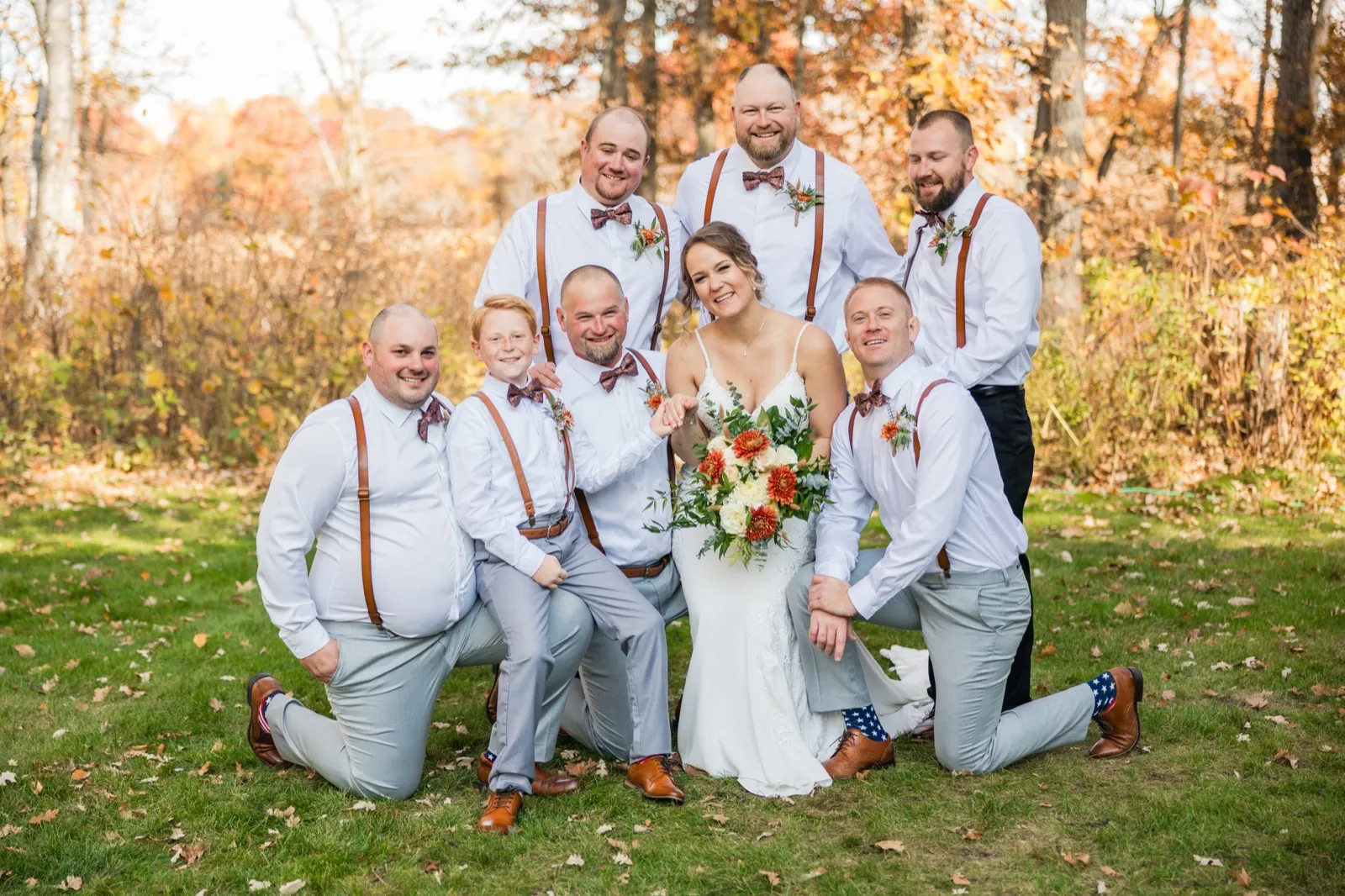 The bride stands at the center of her groom and six groomsmen in suspenders and bow ties, posed on an autumn lawn with golden foliage behind them — Tim Larsen Photography, Brainerd Lakes MN